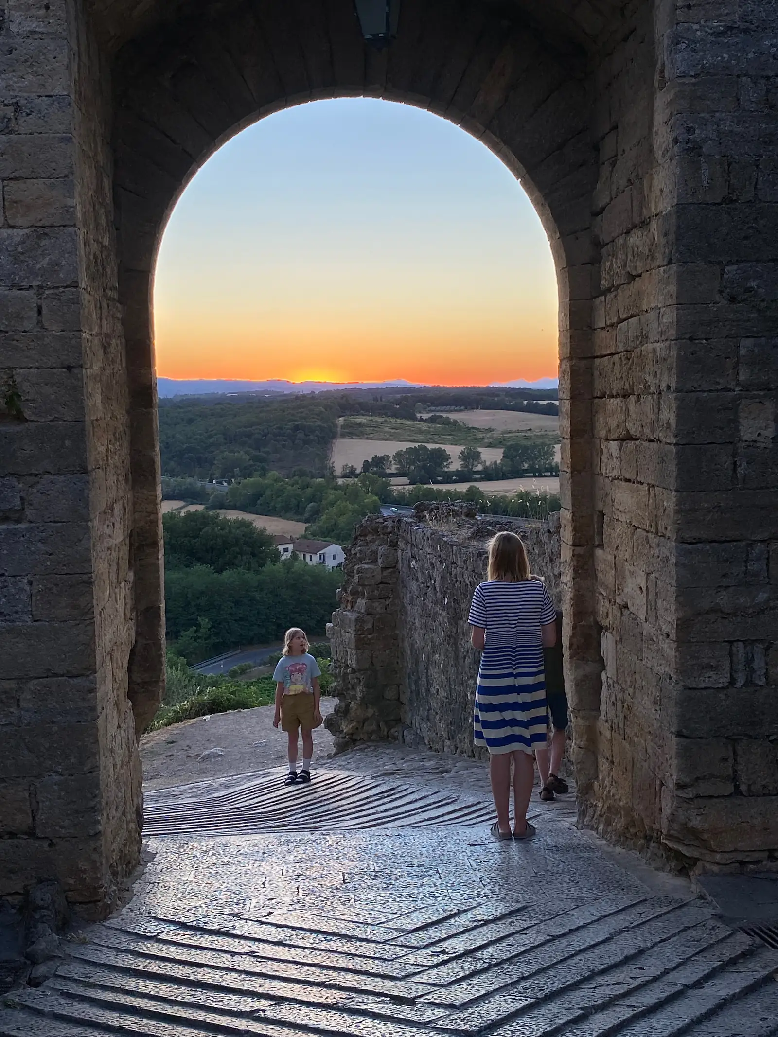 A woman in a striped dress and a child look out from a stone archway at a fiery sunset over the Tuscan countryside below.