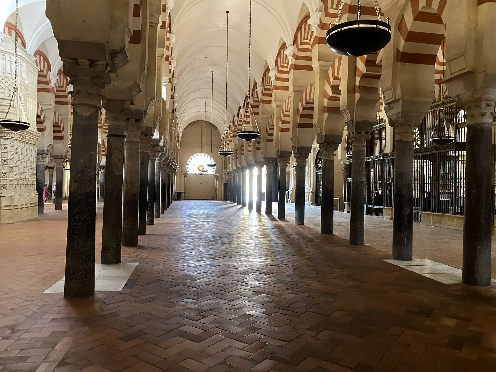 A vast hall of columns and red-and-white striped arches with hanging bowl lamps, sunlight falling in bars across the herringbone brick floor.