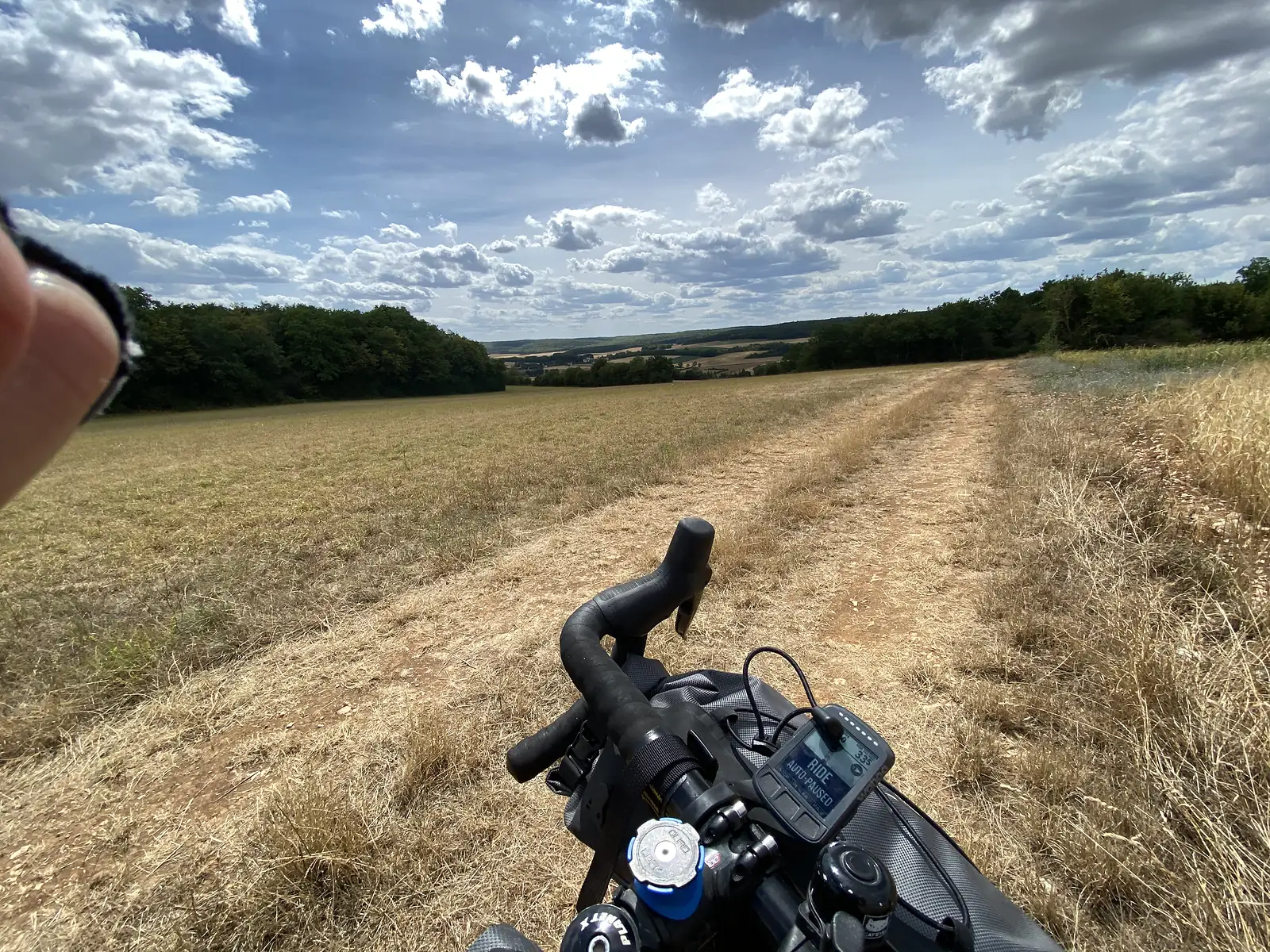 A rutted dirt track crosses a mown field toward a distant belt of trees under a sky of scattered cloud, a rider's fingertip and handlebar bag visible in the foreground.