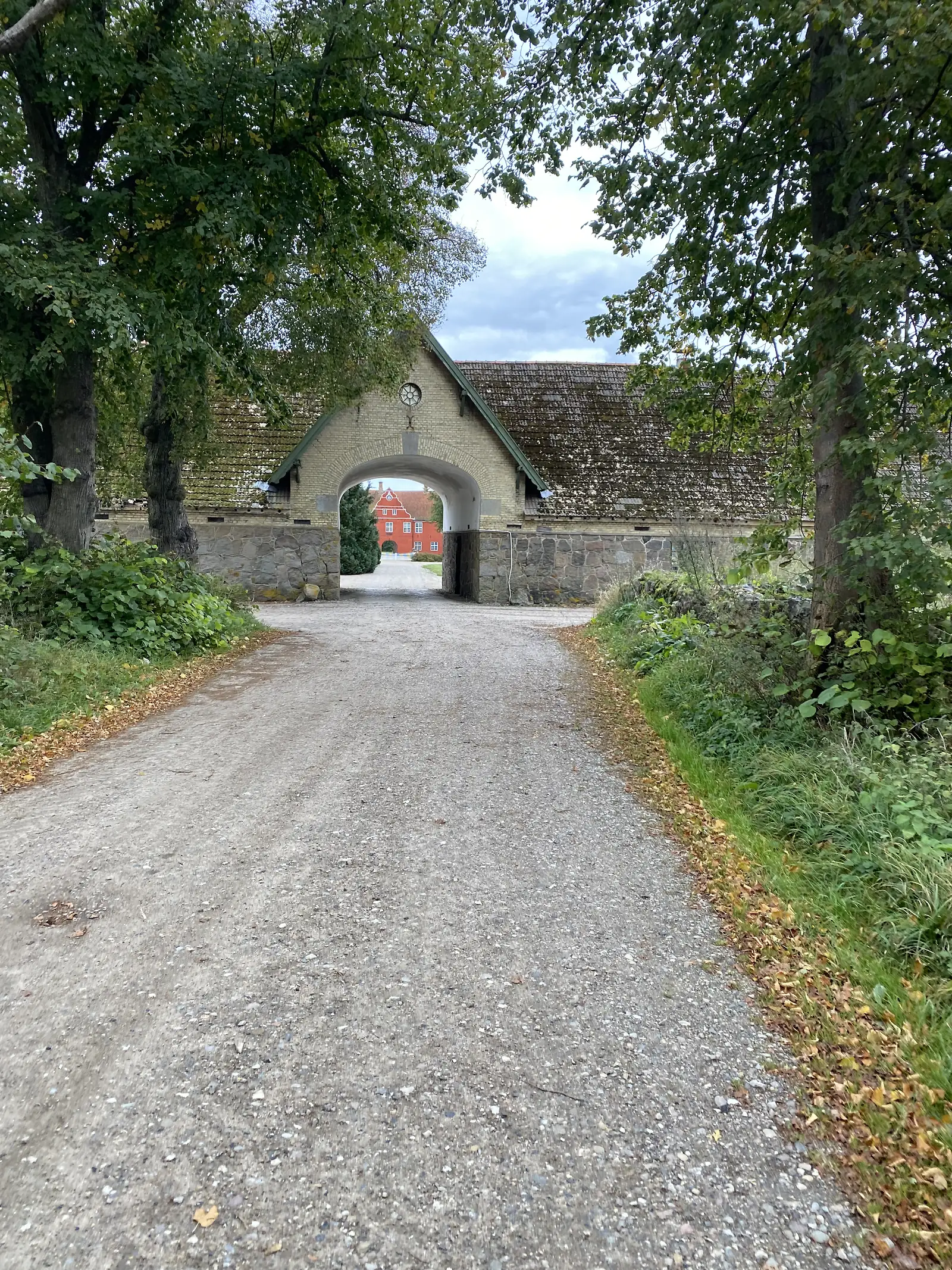 A gravel drive leading to a yellow-brick gatehouse arch in a long moss-roofed farm range, with a red house visible through the arch.