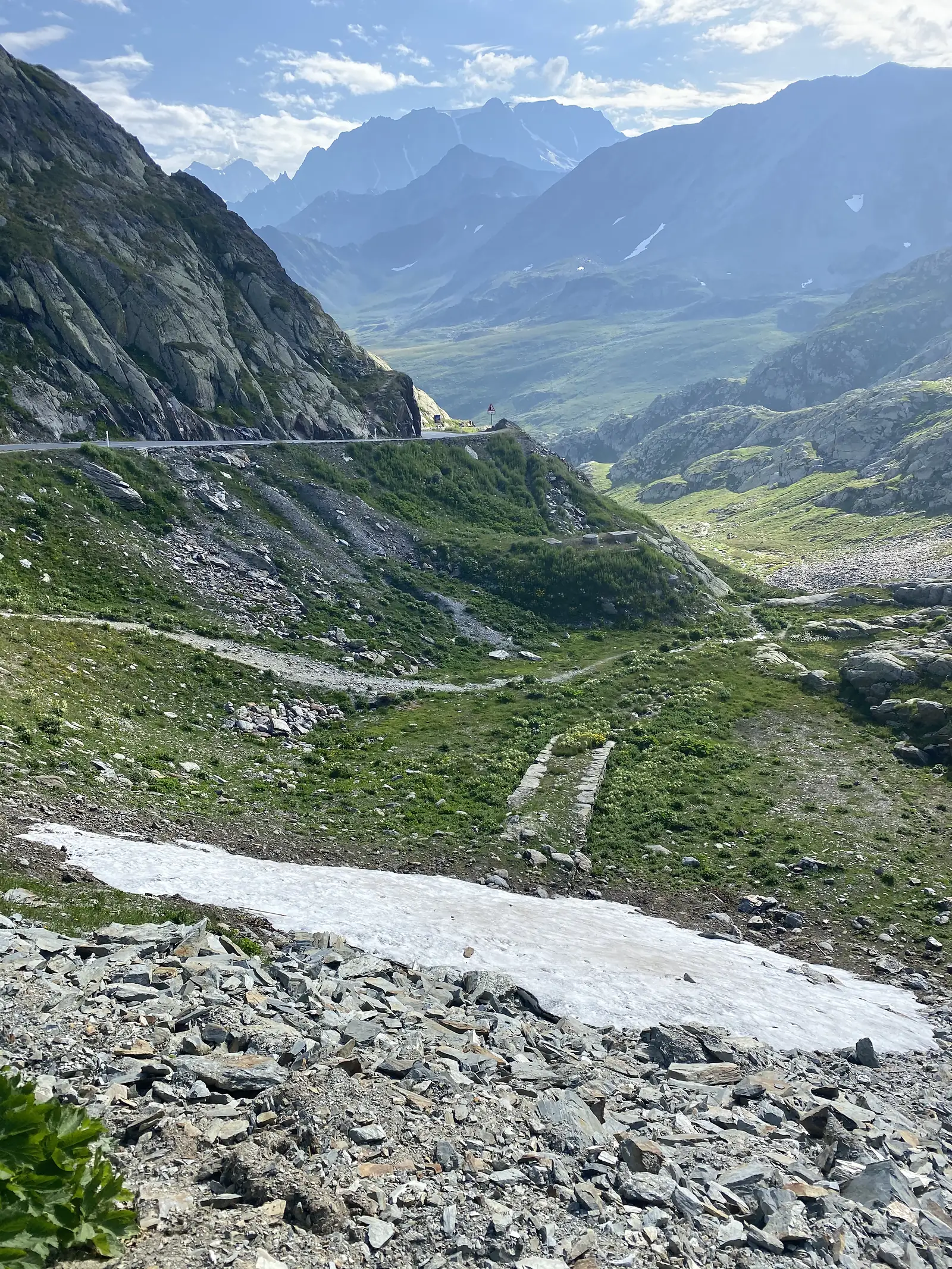 A patch of late-lying snow lies beside a stone-paved path descending through a rocky mountain valley, hazy peaks visible in the distance.
