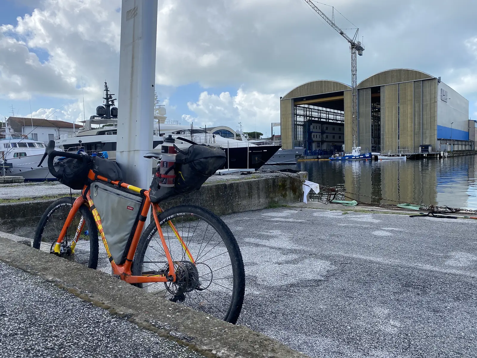 A loaded orange gravel bike leans against a bollard at a marina, with moored motor yachts and a vast boatbuilding shed and crane behind.