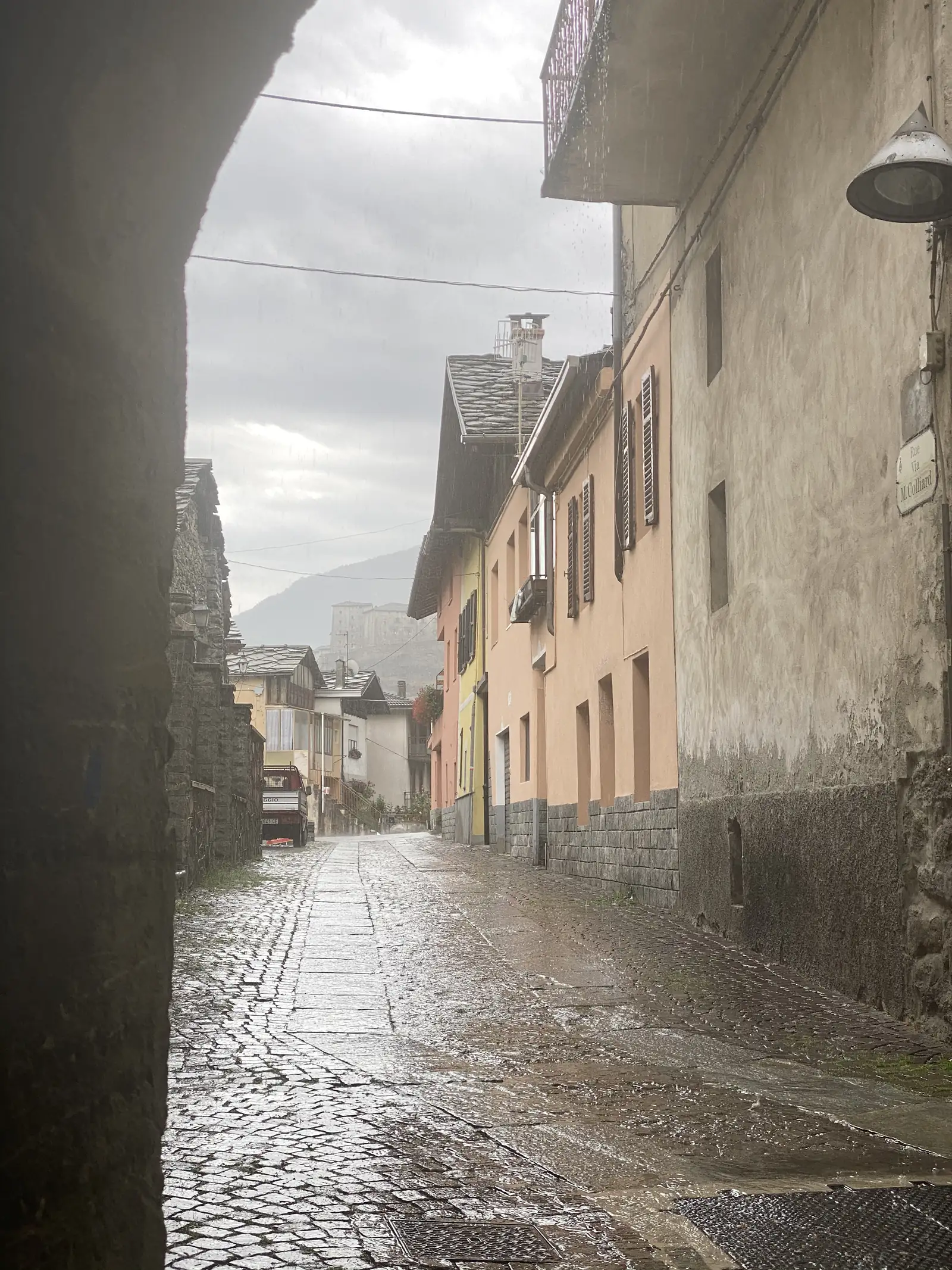 A wet cobbled lane between pastel-coloured village houses, with the pale outline of a hilltop fortress visible in the distance under heavy cloud.