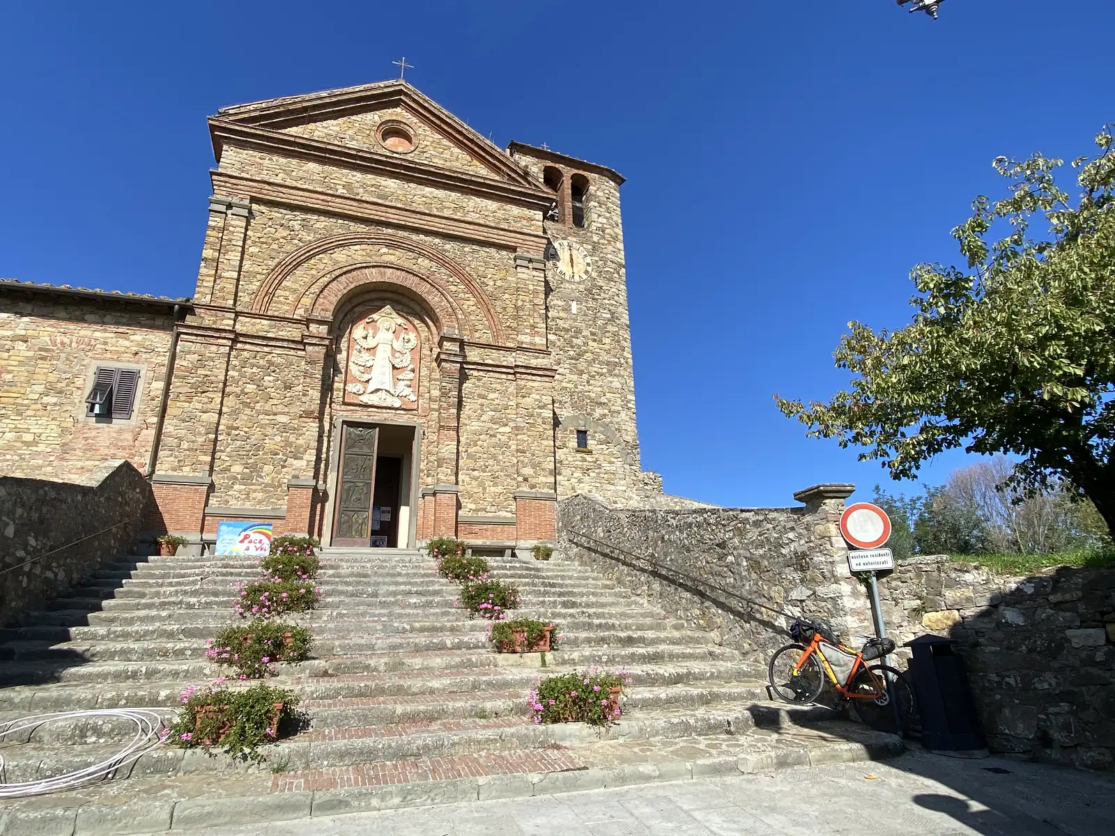 A stone church with a bell tower and clock, its steps lined with pink flowers and a sign reading "Pace a voi", a loaded orange bike parked at the foot of the stairs.