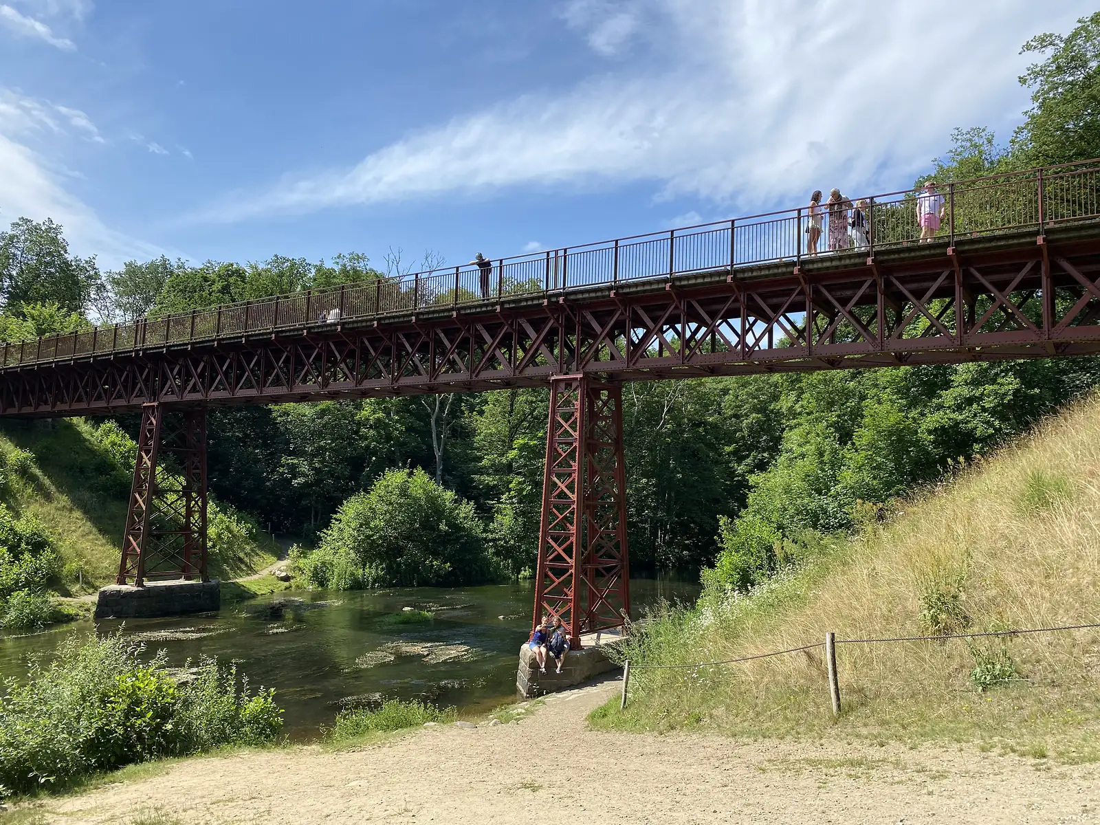 A red-painted steel lattice railway viaduct crossing a wooded river valley, with people on the walkway and two sitting on a pier base by the water.
