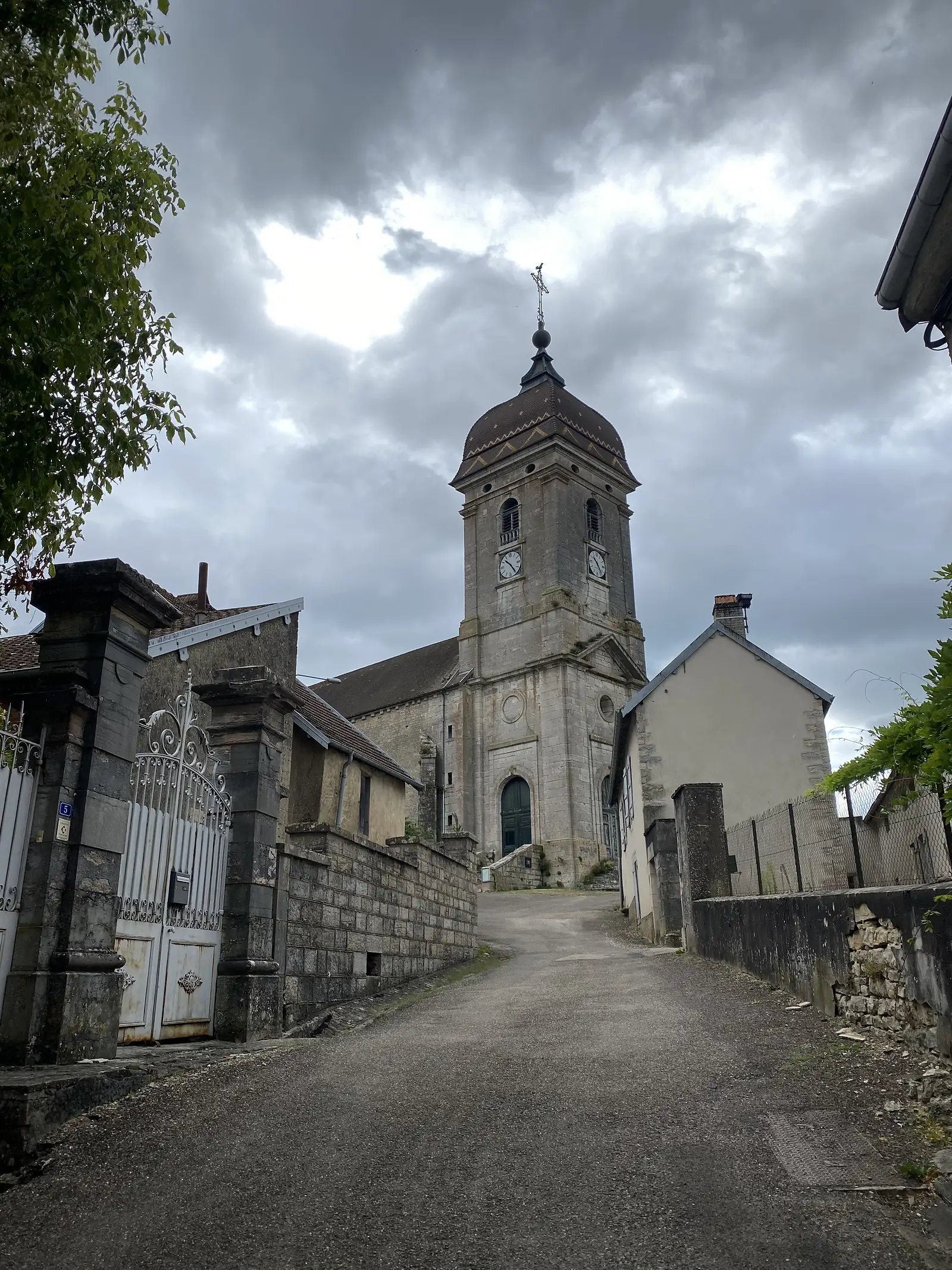 A village church with a domed bell tower and clock faces rises above rooftops at the end of a lane, seen past an old iron gate under a heavy grey sky.