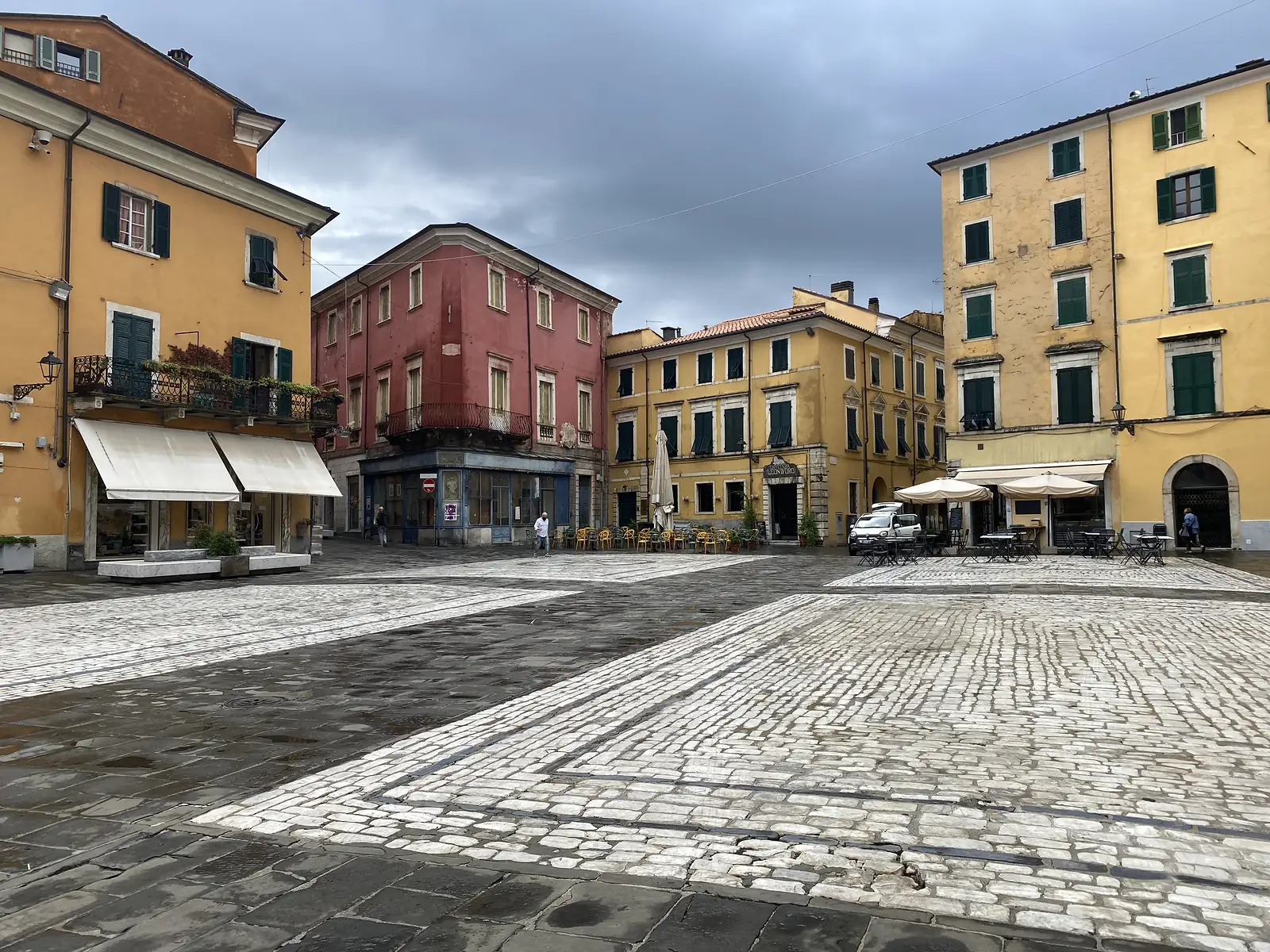 A quiet piazza of ochre, terracotta and dusky-pink buildings with green shutters, its cobbles set with pale marble strips, café tables and umbrellas standing empty under a stormy sky.