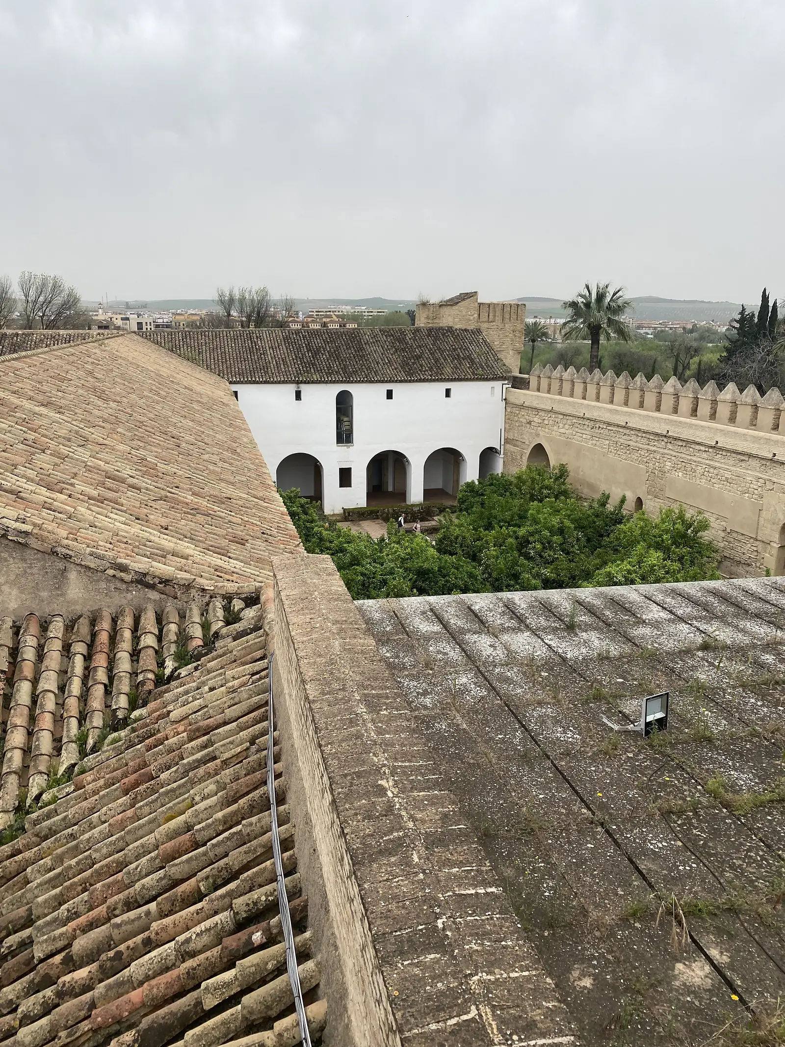 Tiled palace rooftops looking across to a whitewashed arcaded range and a battlemented curtain wall, with orange trees in the garden between.