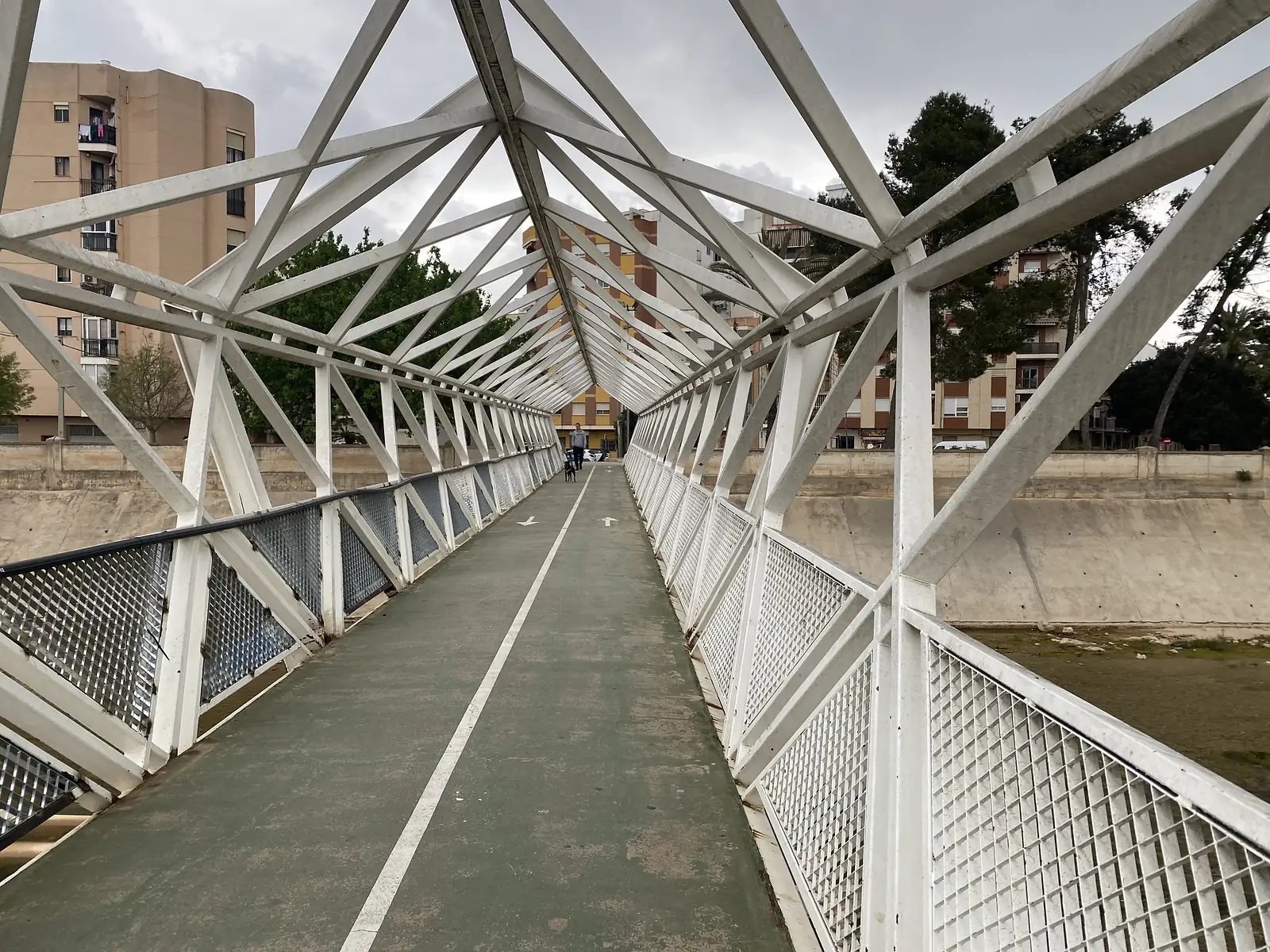 A white steel lattice footbridge carrying a green cycle path over a concrete flood channel between apartment blocks.