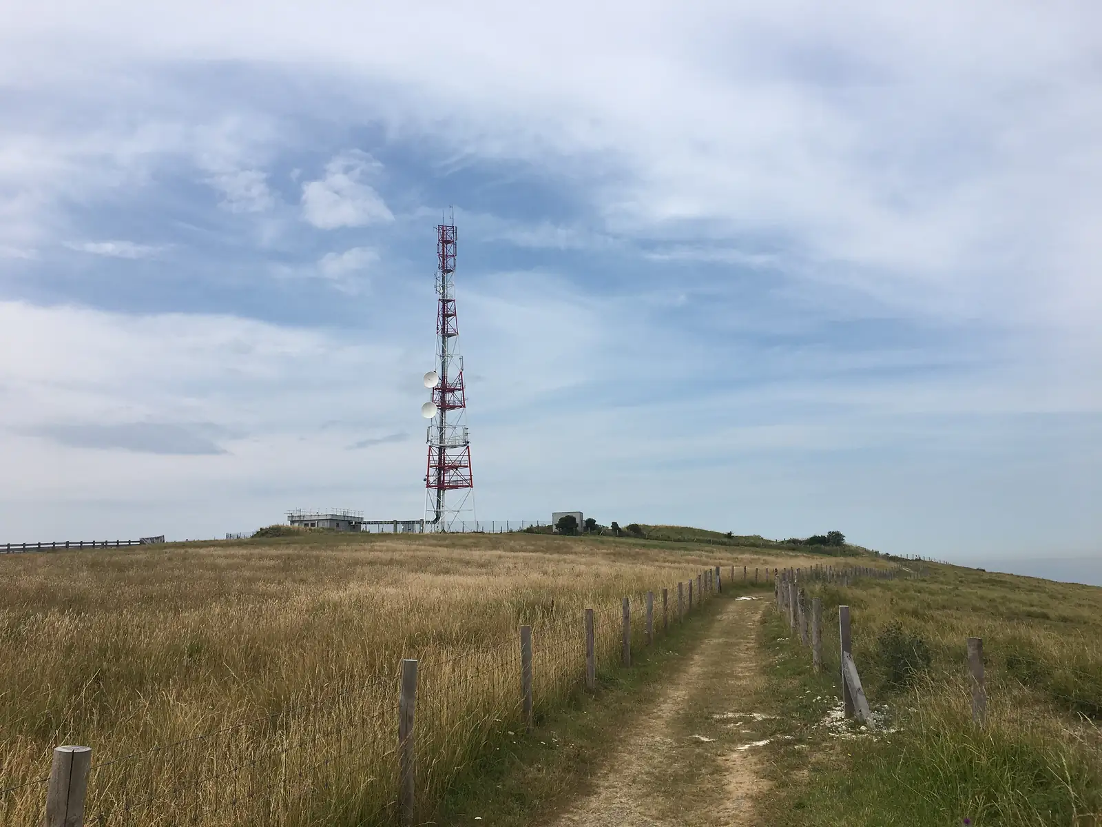 A dirt path bordered by wooden fence posts leading up to a red-and-white telecommunications mast on the brow of a hill.