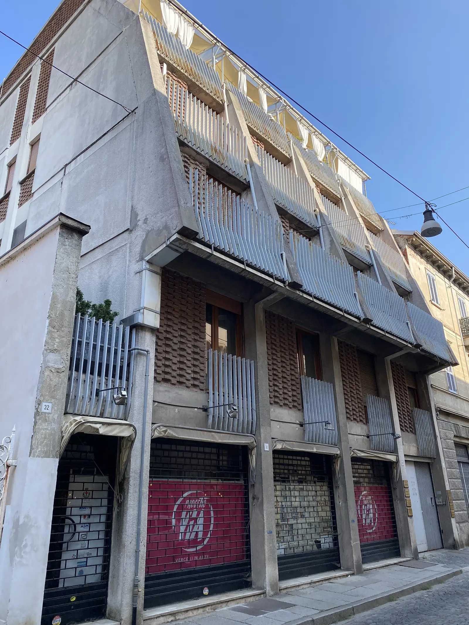 Shuttered apartment balconies above street-level shops on a Vercelli street, a red banner reading 'VIA' hanging over one of the shopfronts.