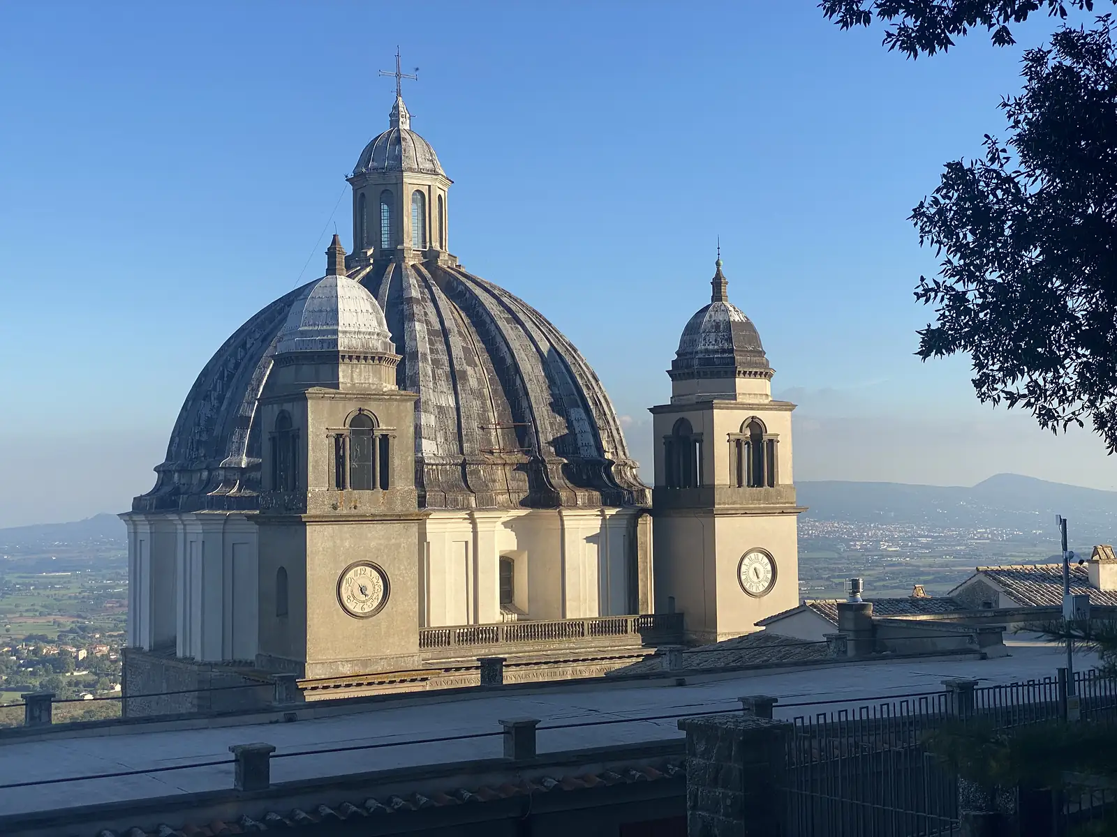 A large church dome flanked by twin clock towers rises above the rooftops of a hilltop town, with a hazy plain stretching to distant hills.