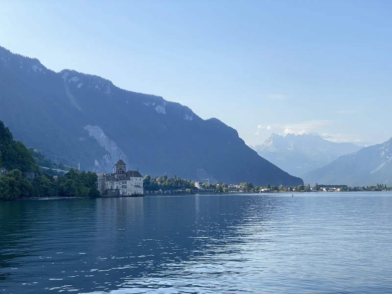 A turreted lakeside castle sits at the water's edge beneath steep wooded mountains, a lone kayaker visible far out on the calm lake.