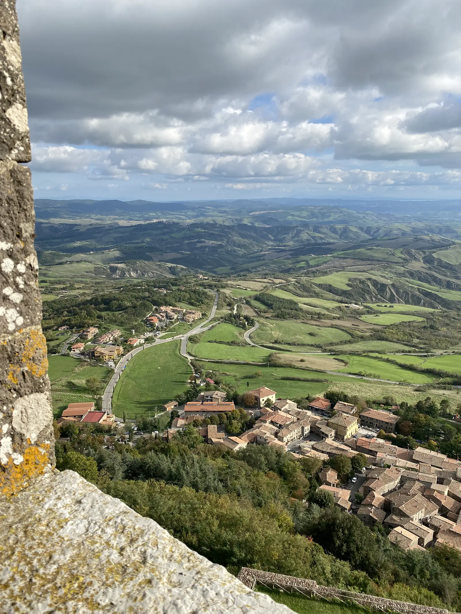 A bird's-eye view over a lichen-covered stone parapet down onto a hilltop town's terracotta rooftops, with a green valley and layered hills stretching to the horizon.