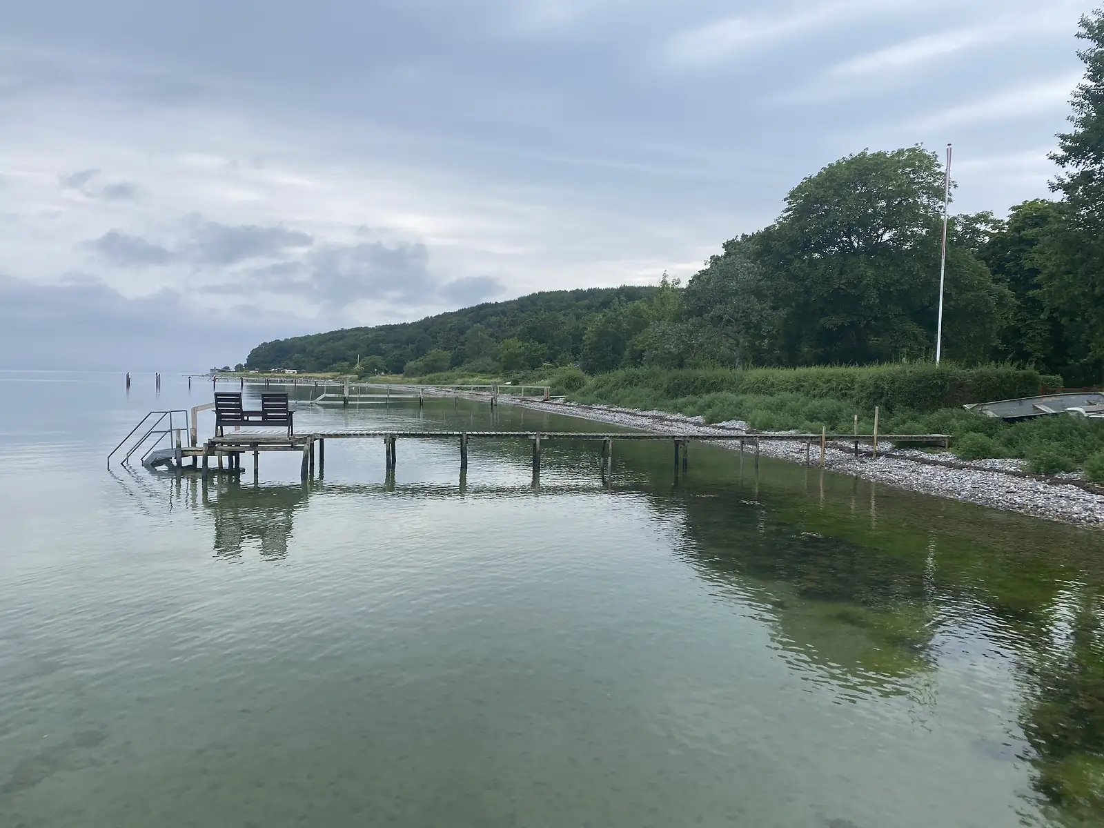 A long timber jetty with two deckchairs at its end reaching into a glassy calm sea, with a wooded shore alongside.