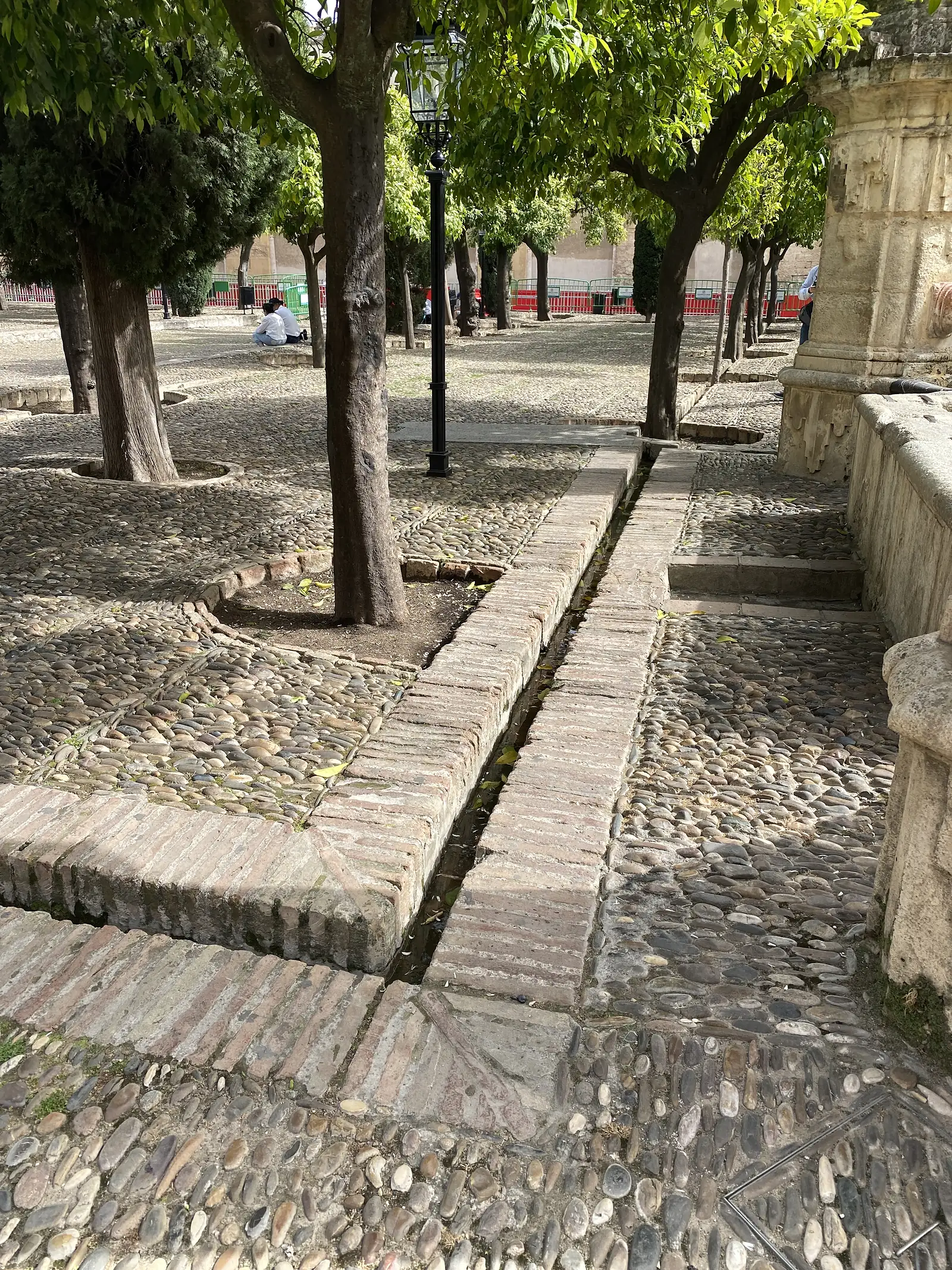 A cobbled courtyard of orange trees with a narrow brick water channel running between them and a black lamp post at the centre.