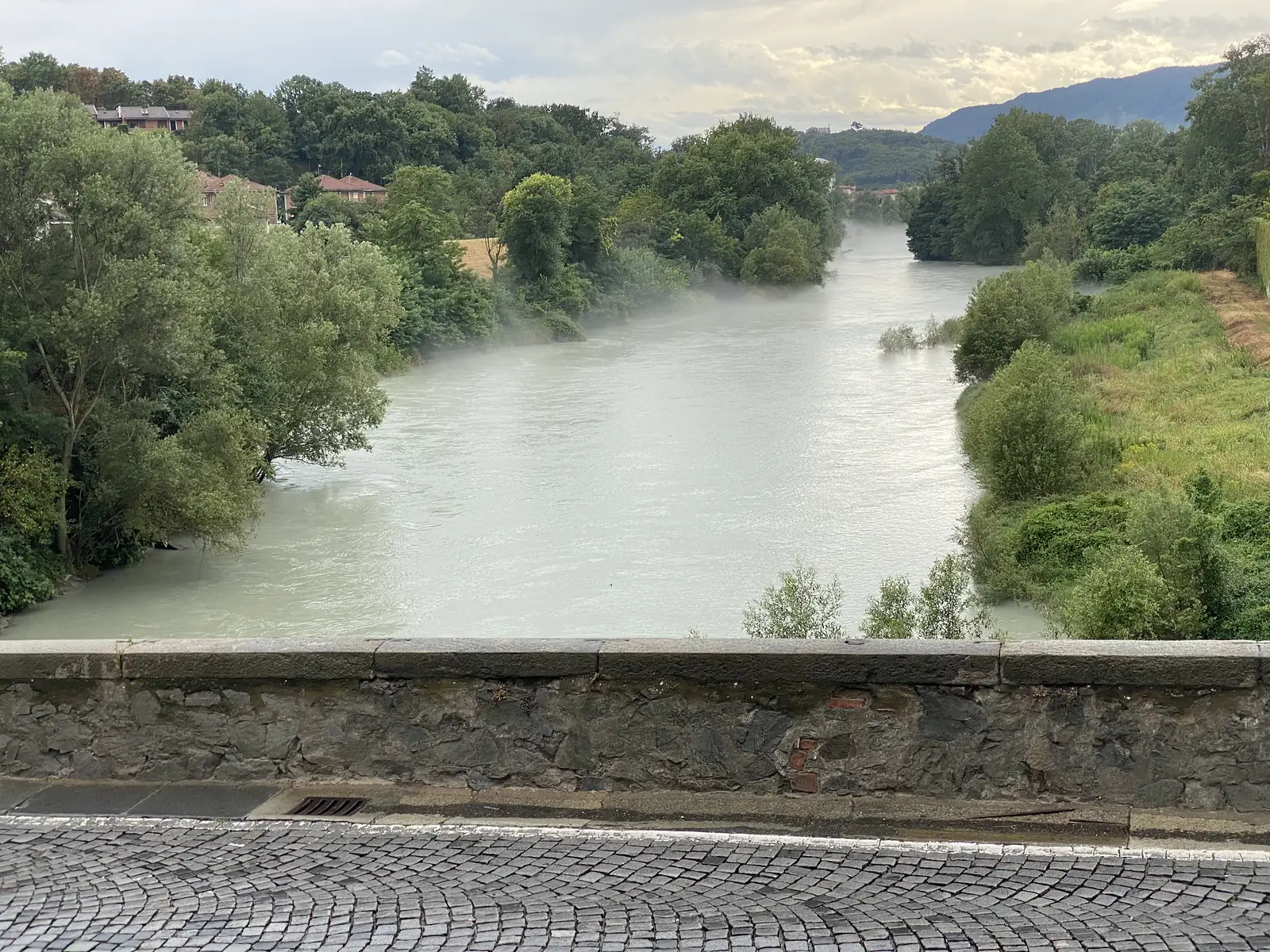 A wide, mist-covered river flows between wooded banks below a stone parapet, with hills and a distant hilltop building visible through the haze.