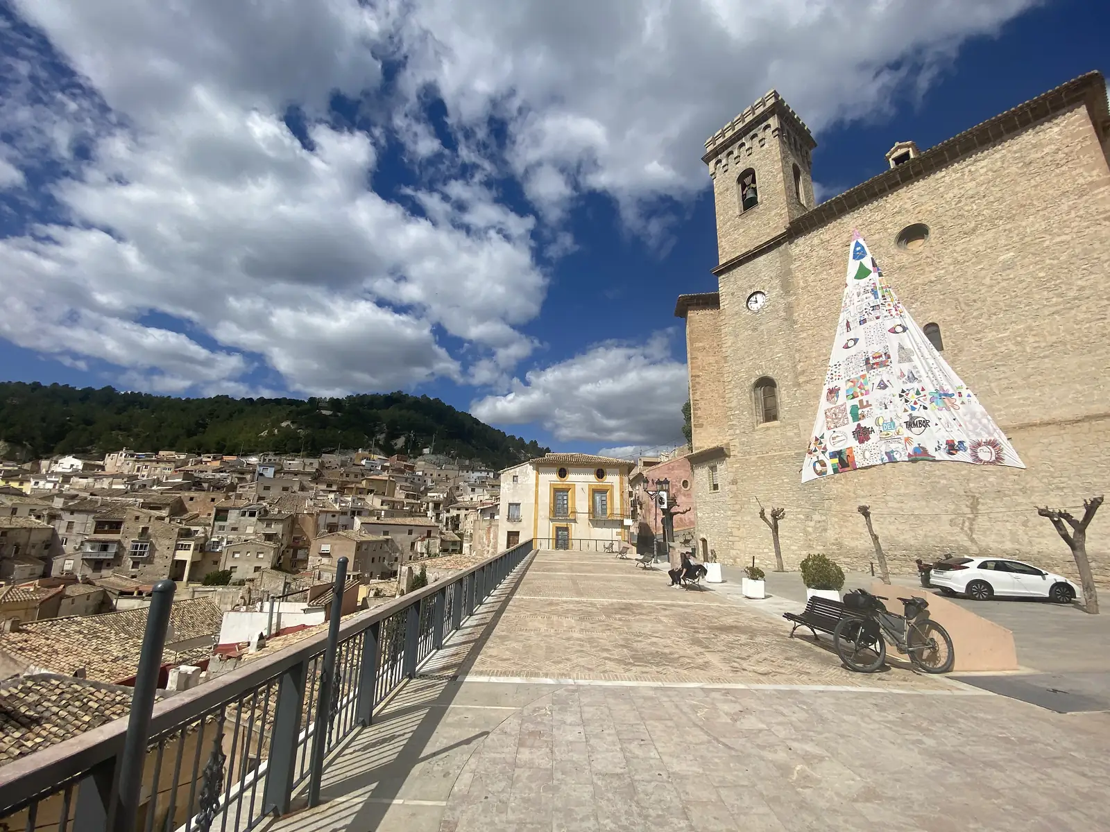 A loaded bike propped by a bench on a wide brick terrace beside a stone church, a huge painted banner reading "FIESTA DEL TAMBOR" hung down its wall above the town's rooftops.