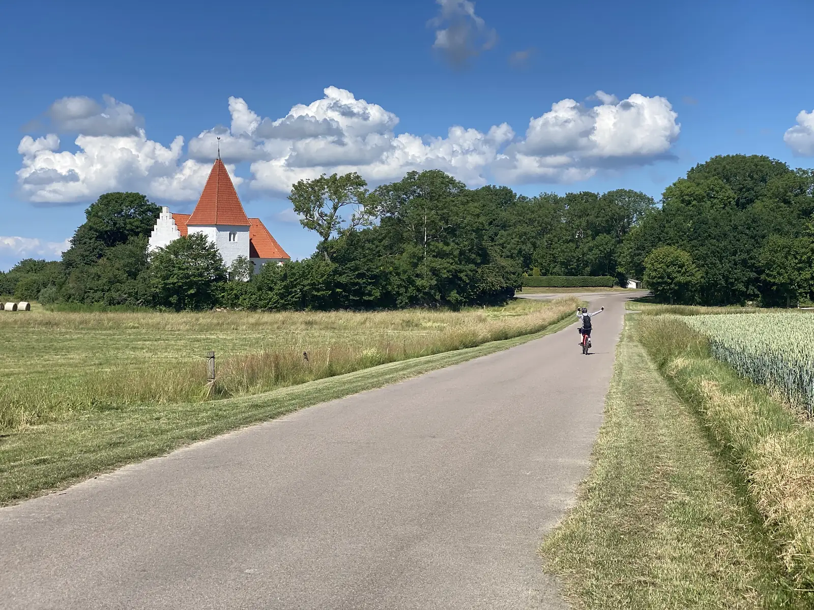 A cyclist riding away along an empty country road past a white stepped-gable church with a red spire, hay meadow and cereal either side.