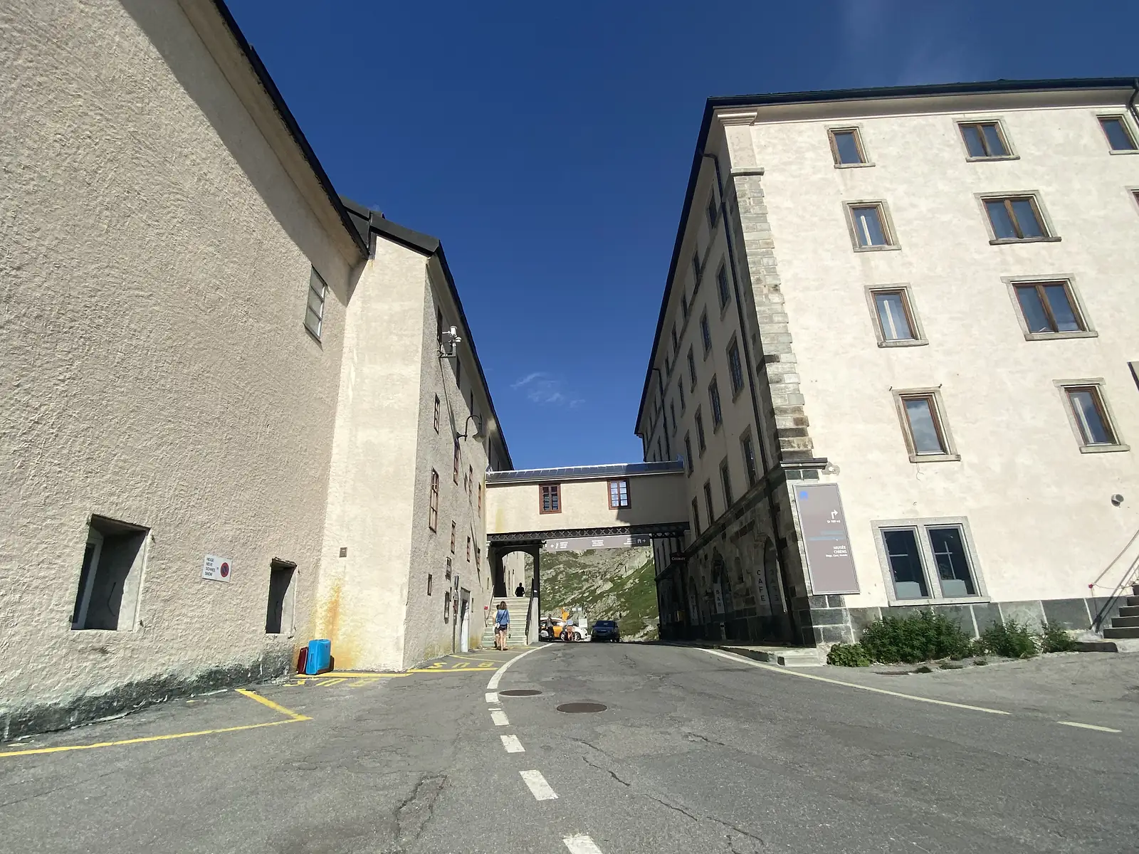 A covered walkway links two tall stone buildings at the mountain pass, with a 'Musée Chiens' sign and open hillside visible through the gap.