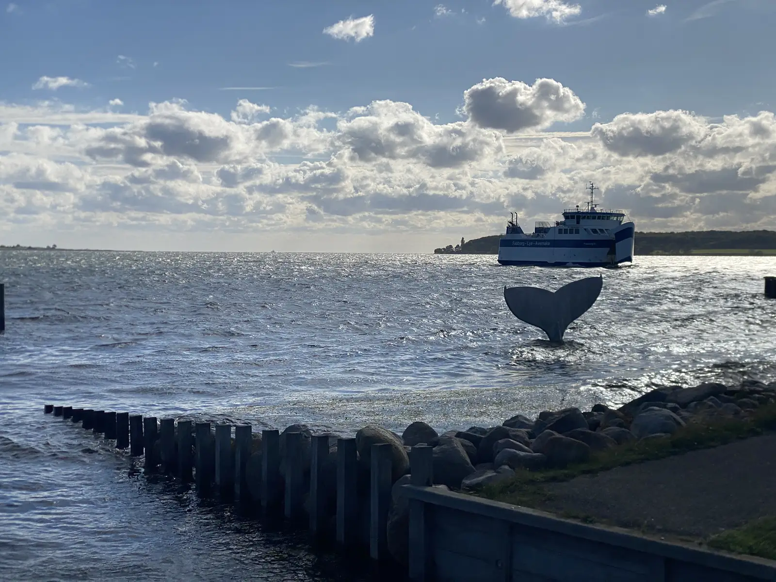 A small ferry lettered "Faaborg - Lyø - Avernakø" crossing a bright sea beyond a whale-tail sculpture rising from the shallows.