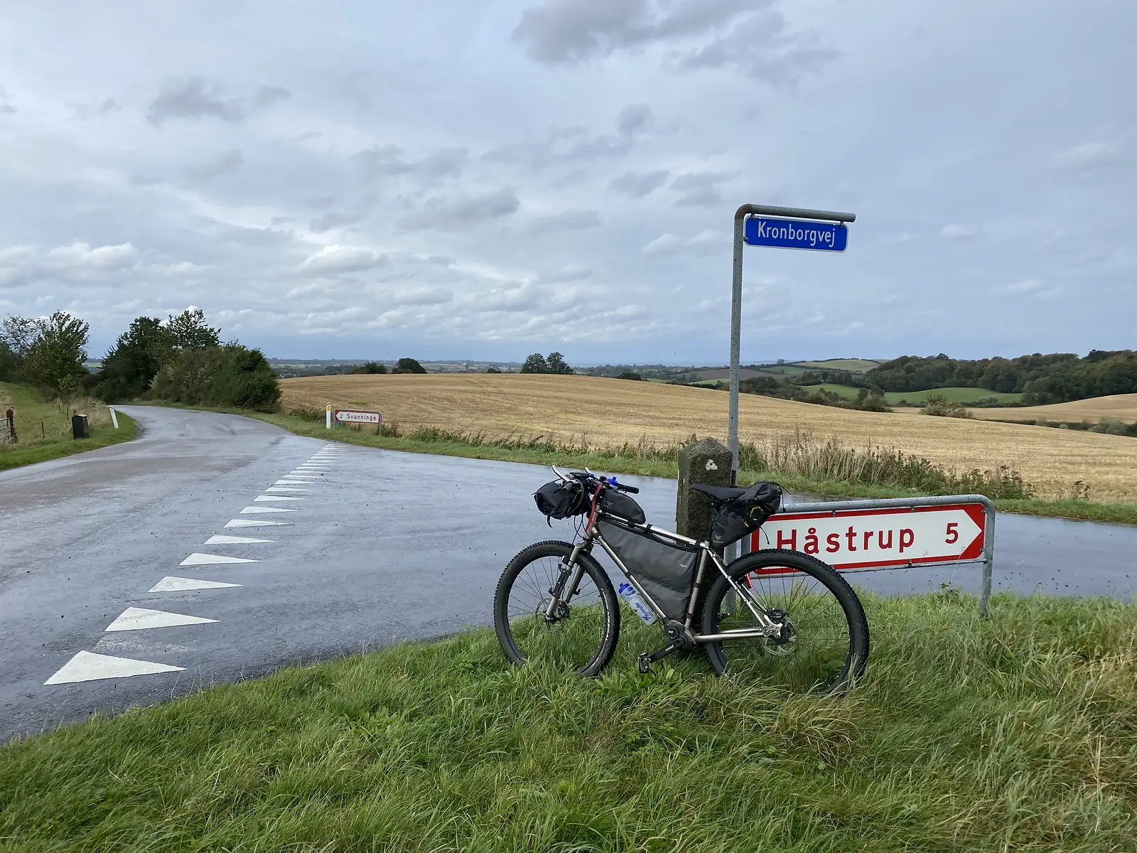 A loaded bikepacking bike leaning on a red-and-white direction sign reading "Håstrup 5" at a wet crossroads, with harvested fields running down to the sea.