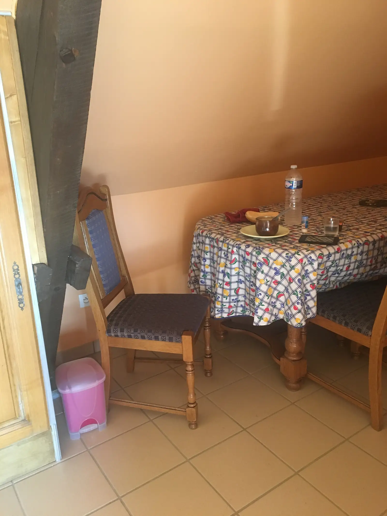 A gite dining table laid with a fruit-patterned oilcloth, water bottle, phone and bread, flanked by wooden chairs with blue upholstered seats.