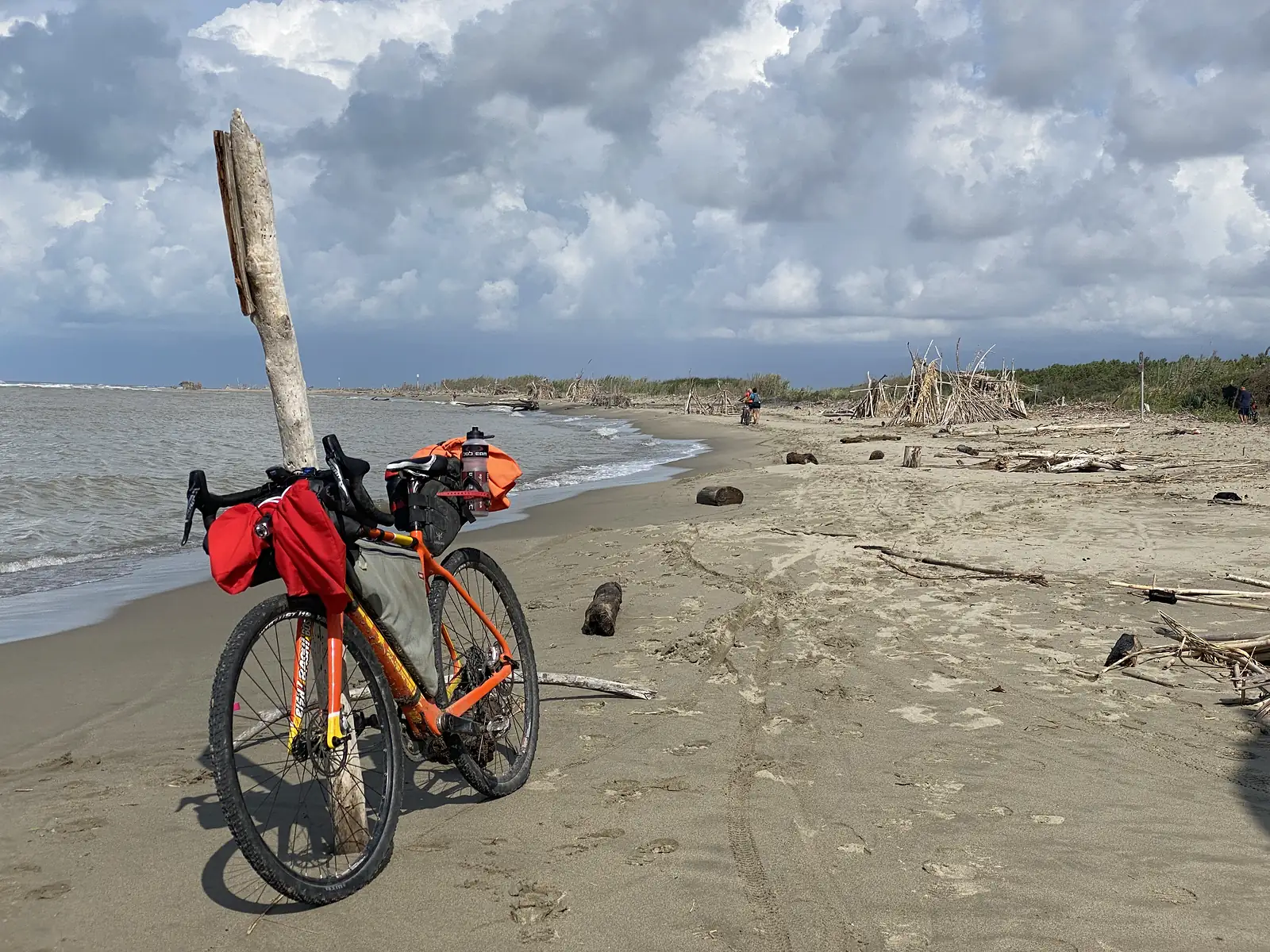 The loaded orange bike resting against a weathered wooden post on the beach, with driftwood shelters and other cyclists in the distance.
