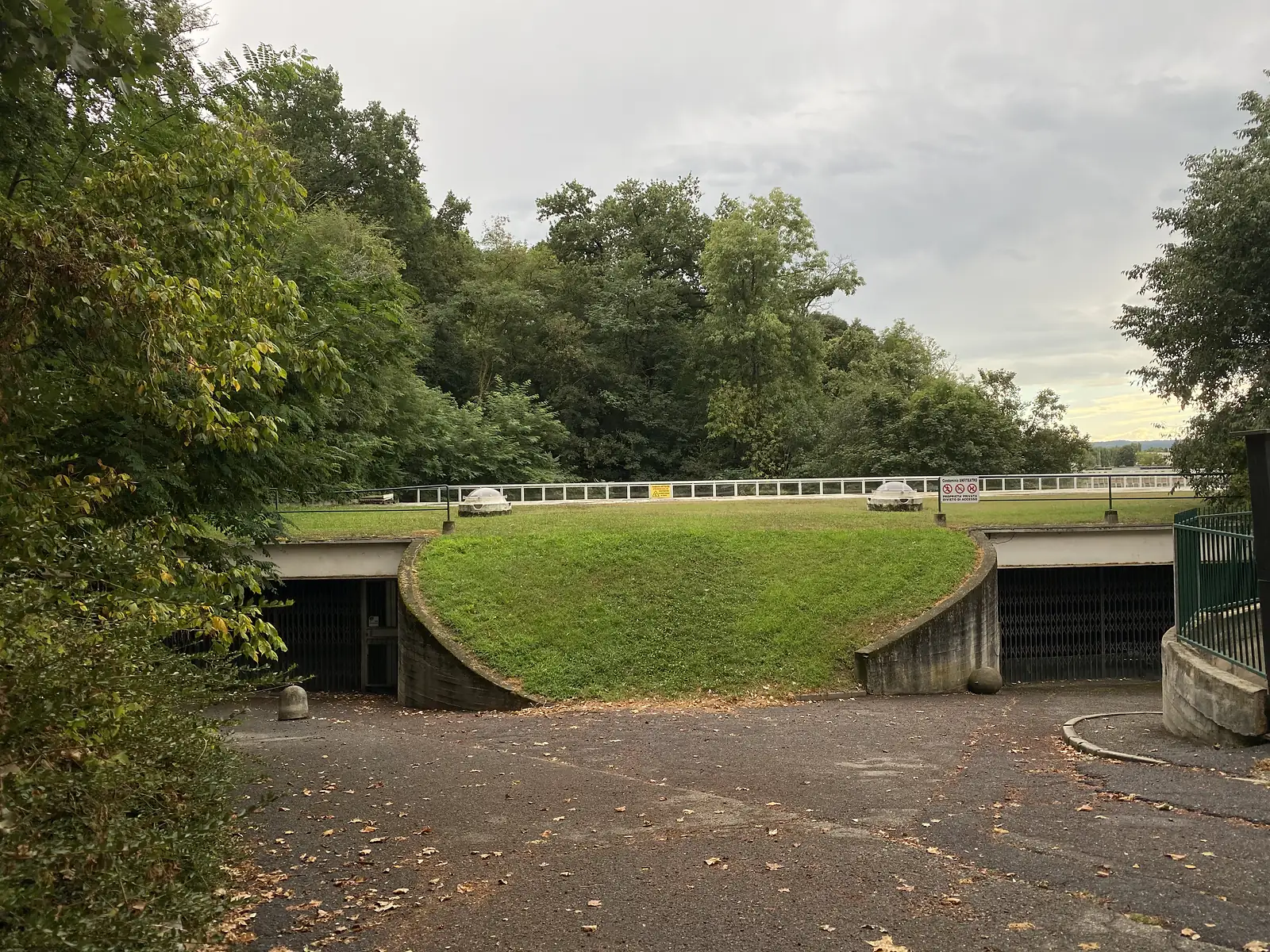 A grassed, sunken courtyard ringed by a white balustrade, domed rooflights set into the lawn above gated tunnel entrances leading underground on either side.