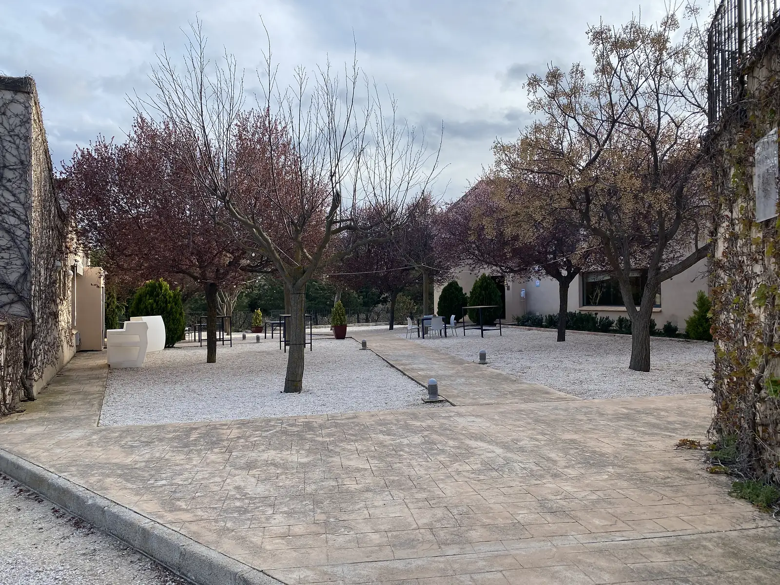 A gravelled hotel courtyard with young trees, festoon lights and bar tables, under a grey evening sky.
