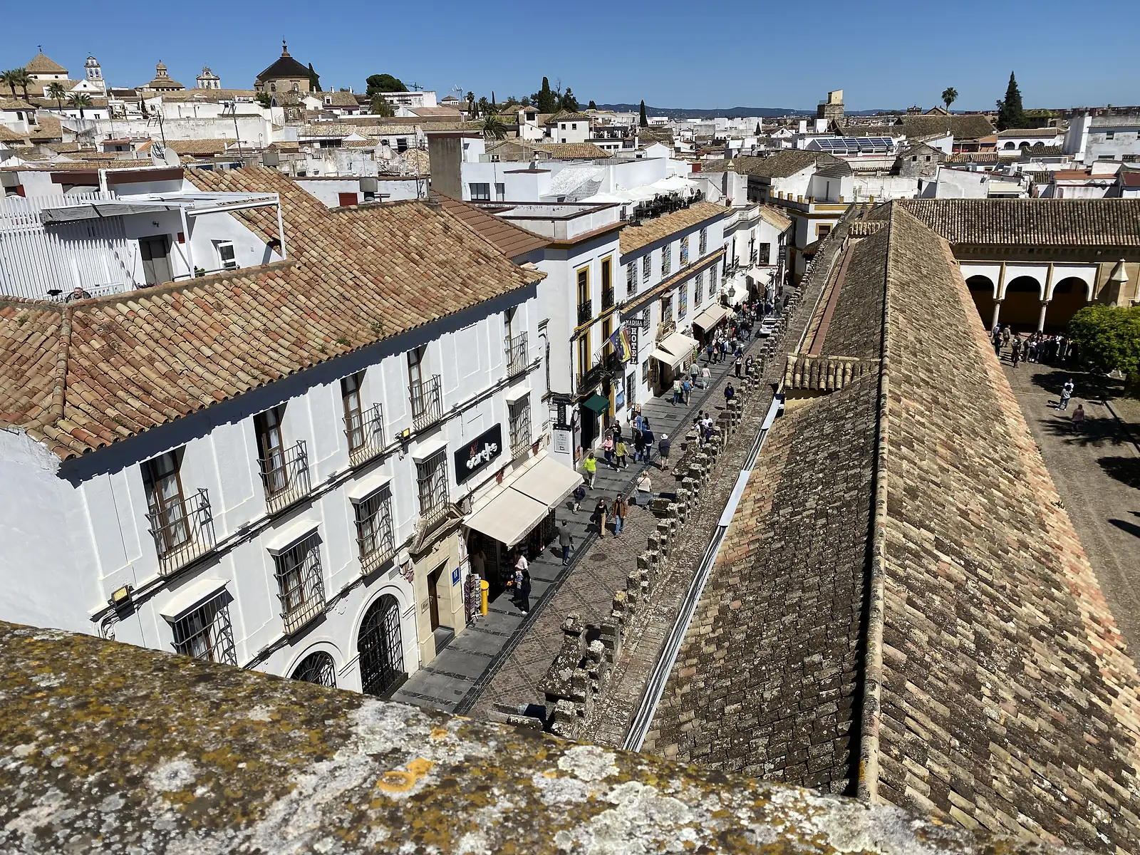 Looking down from a rooftop onto a busy pedestrian street of shops and awnings, with the white old town and a courtyard of arches beyond.