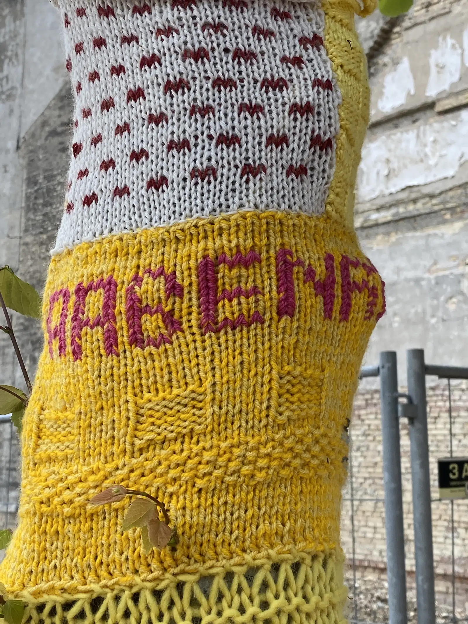 A tree trunk wrapped in knitted yarn-bombing, white with red hearts above a lettered yellow band, against a brick and concrete wall.
