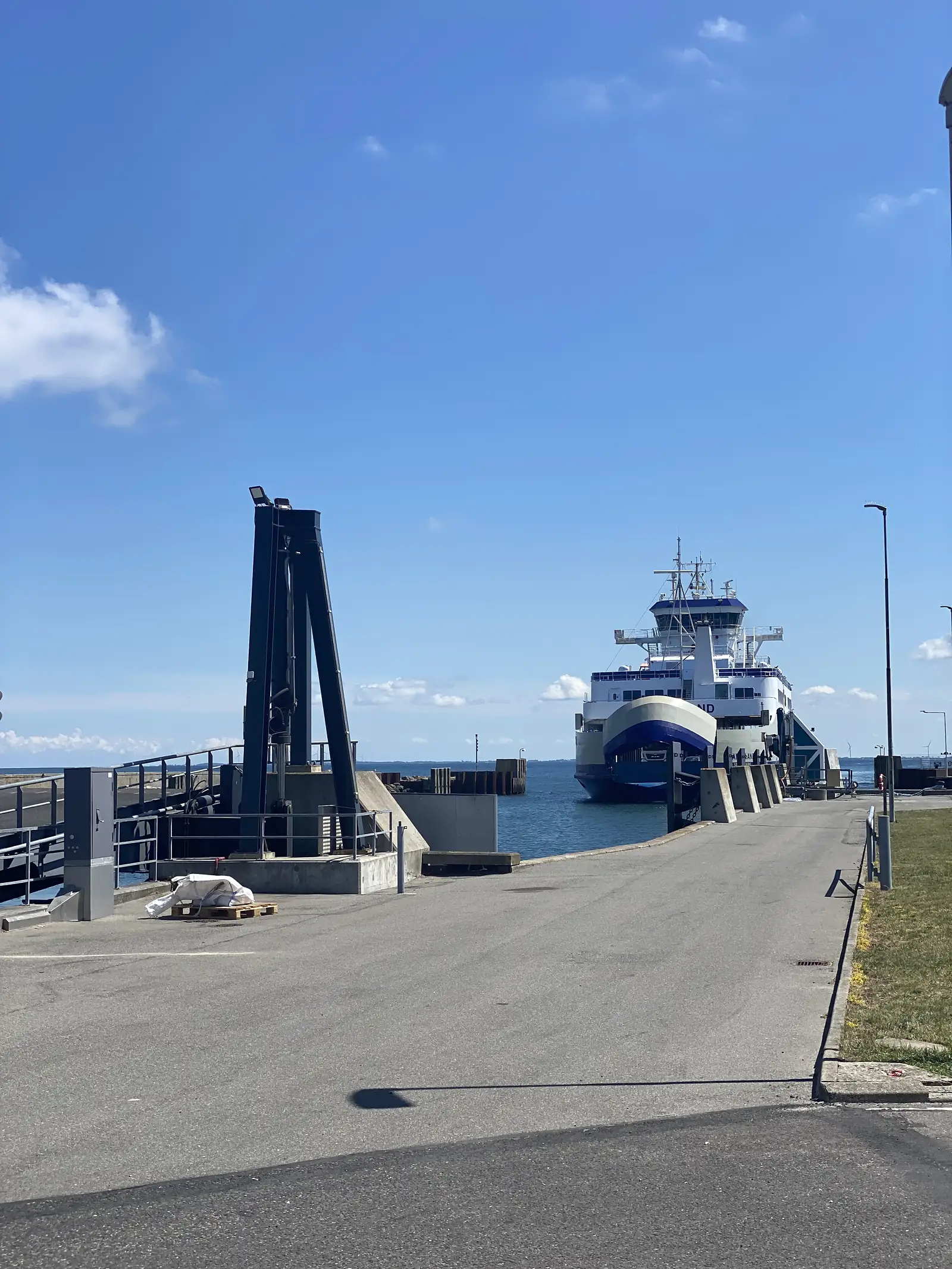 A blue-and-white ferry berthed at a concrete linkspan under a clear blue sky, with a black loading gantry on the quay.