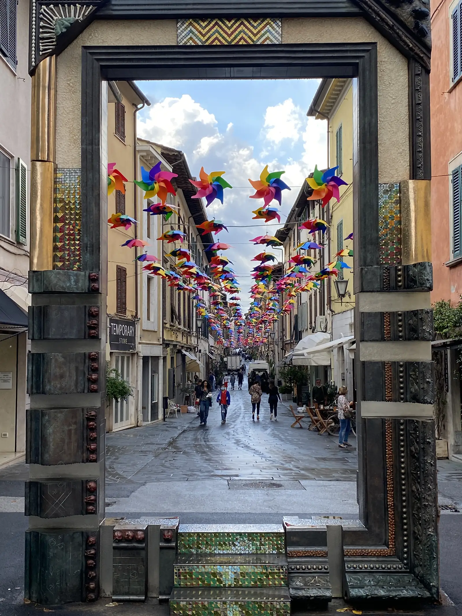 Rows of colourful pinwheels strung across a narrow Tuscan street flutter above shopfronts, including one marked 'Temporary Store', as pedestrians stroll beneath them.