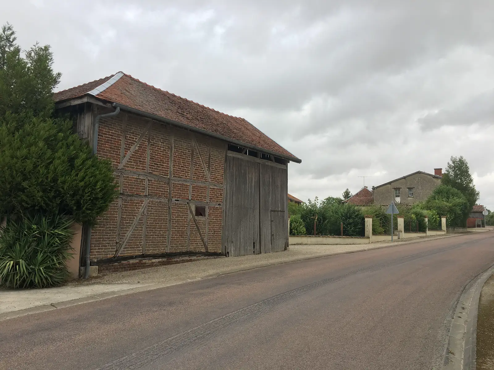 A half-timbered brick barn with weathered wooden double doors and a tiled roof stands beside a quiet village street.