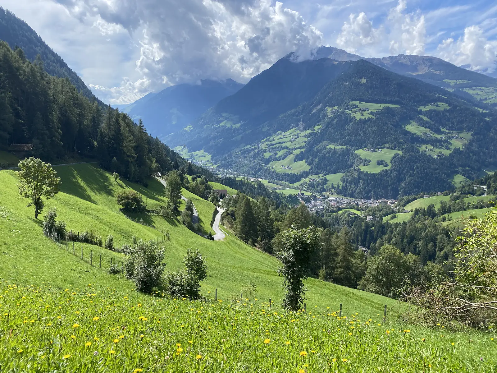 Flowery hay meadows and a hairpin lane drop away to a village on the floor of a deep green valley, with cloud building over the ridges.