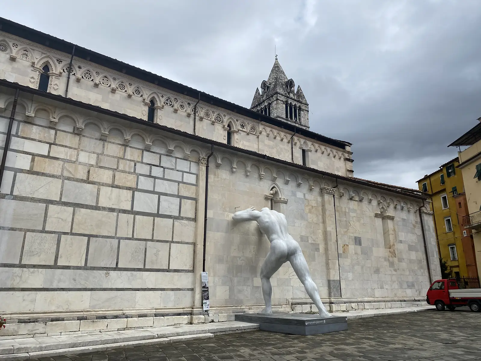 The marble side wall and bell tower of a church, with a stark white sculpture of a crouching male nude figure standing in the piazza in front and a small red truck parked nearby.