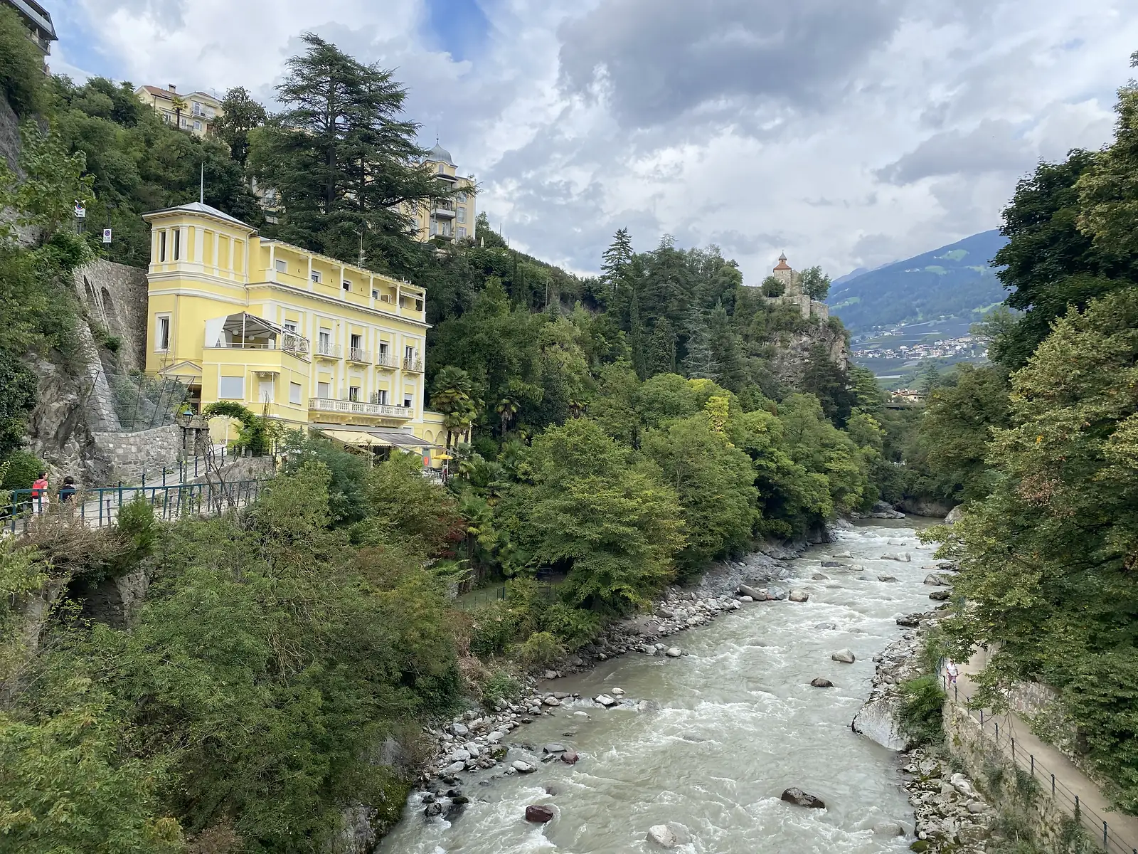 A yellow villa with a colonnaded top floor stands above a milky, boulder-strewn river running through a wooded gorge, a small castle on the crag beyond.