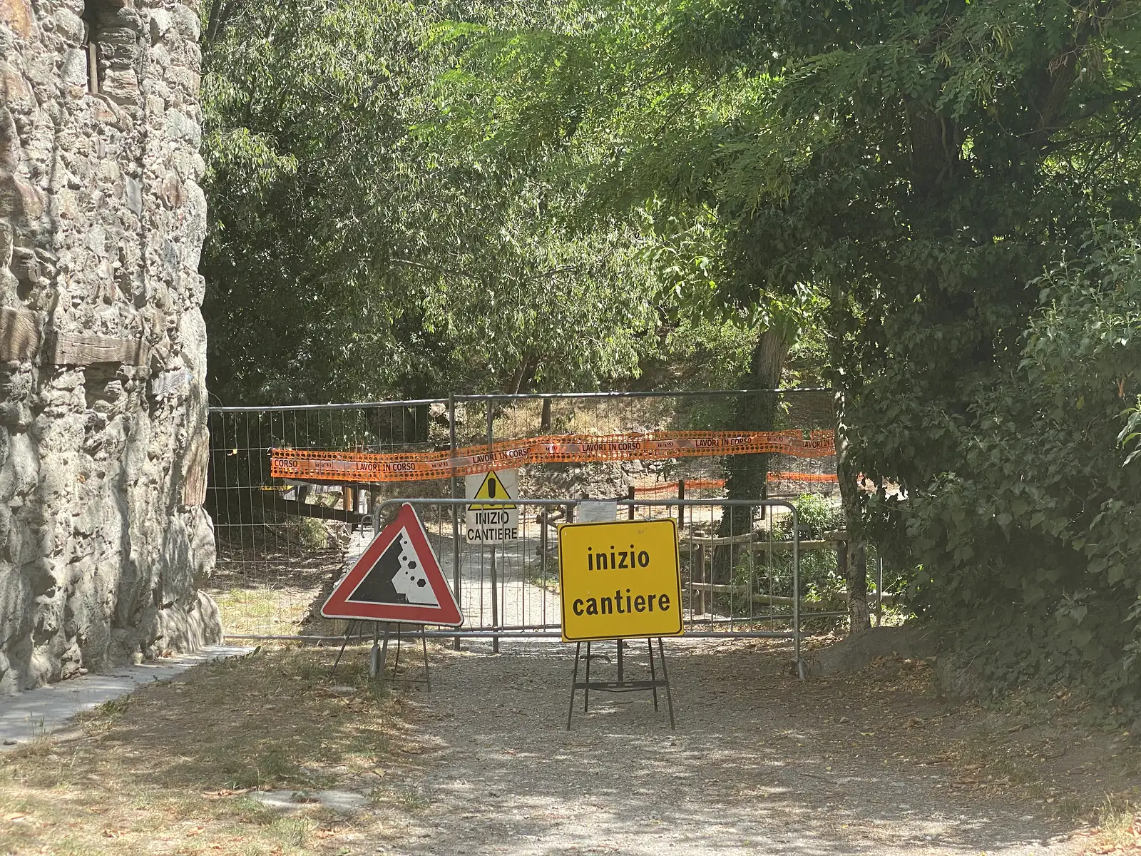 A metal barrier hung with orange 'lavori in corso' tape and a yellow 'inizio cantiere' sign blocks a wooded path beside an old stone wall, warning of a landslide hazard.