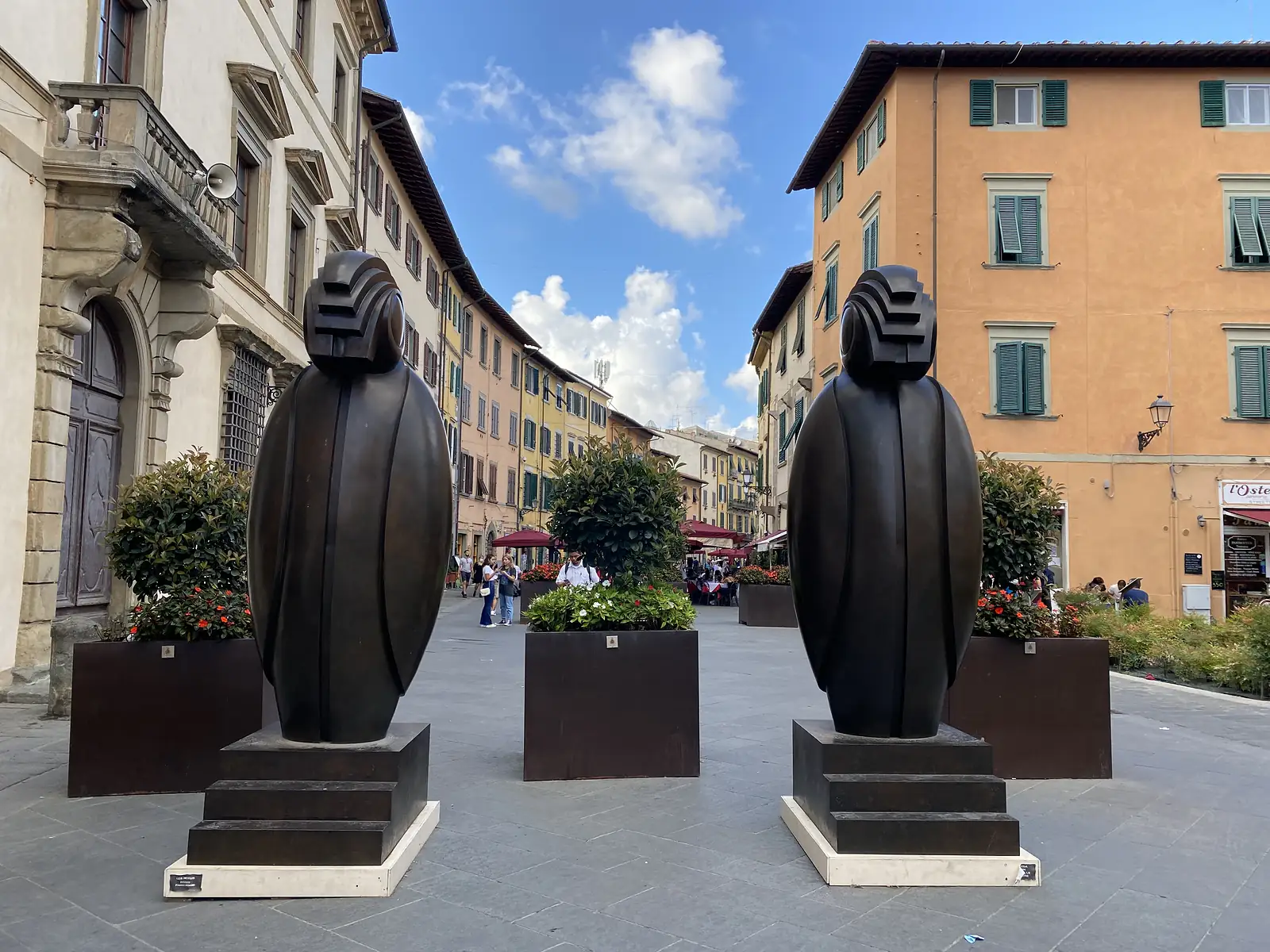 Two tall bronze sculptures of stylised hooded figures stand in a paved Pisan street lined with pastel-coloured buildings and outdoor cafés.