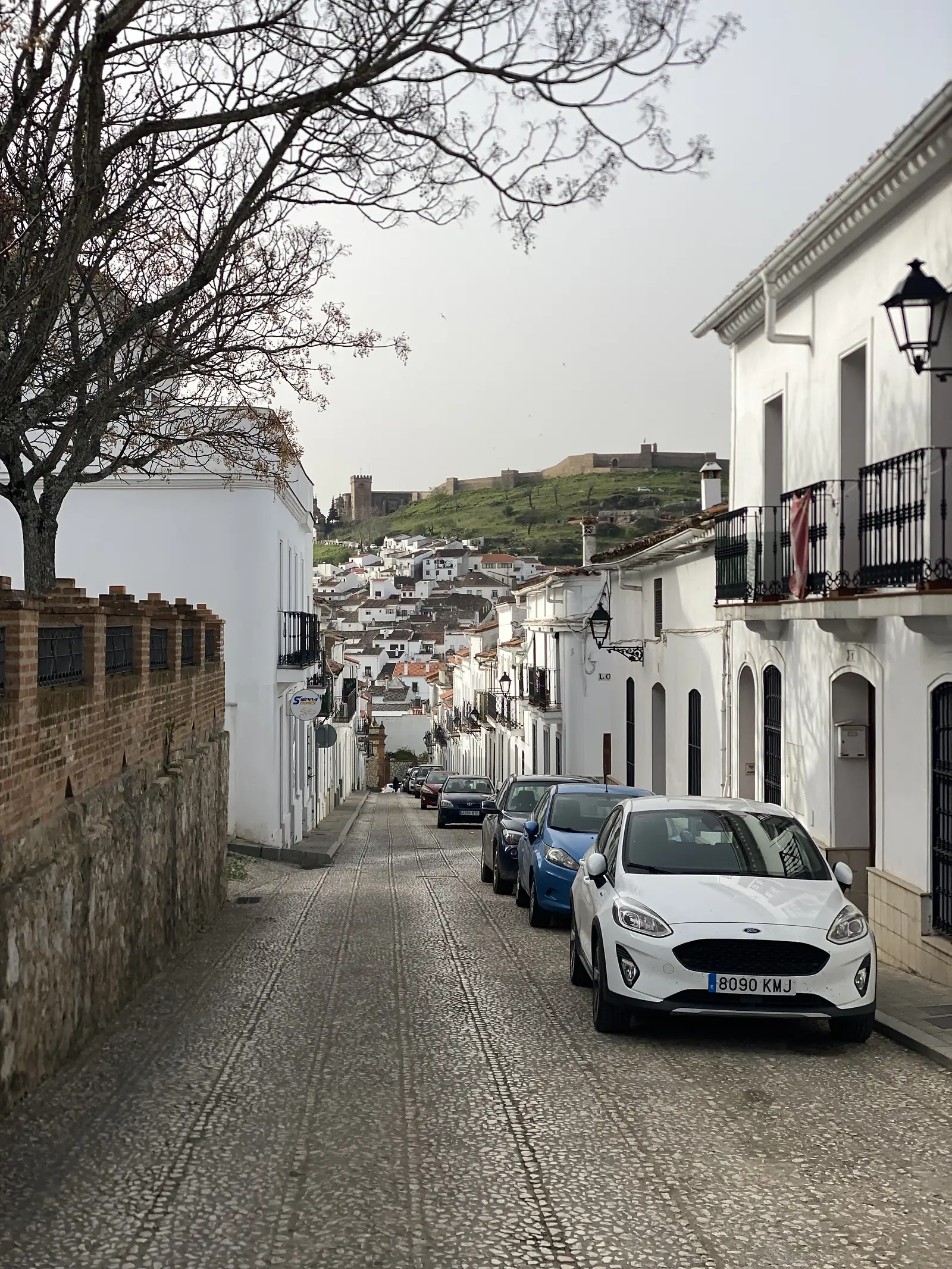A cobbled street of parked cars and whitewashed houses dropping away towards a hillside of white roofs, with a long castle wall on the ridge above.