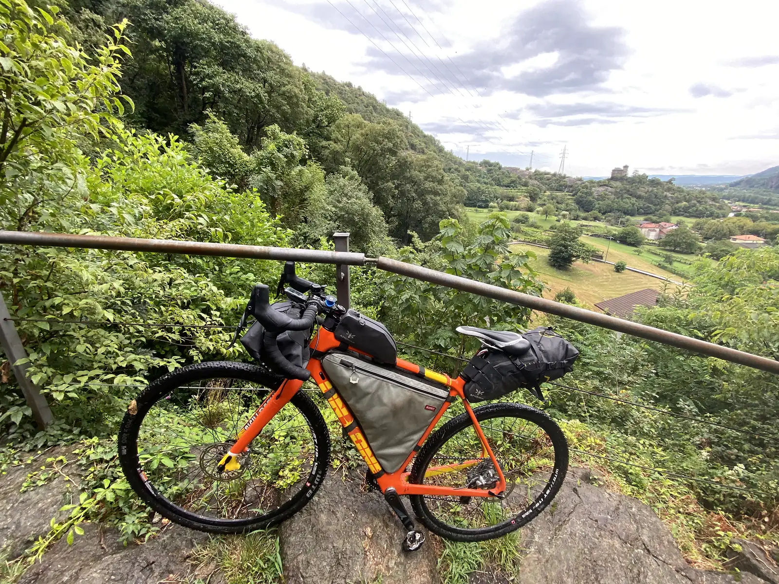 A loaded orange gravel bike rests against a metal railing at a hillside viewpoint, overlooking wooded slopes and a green valley with farm buildings below.