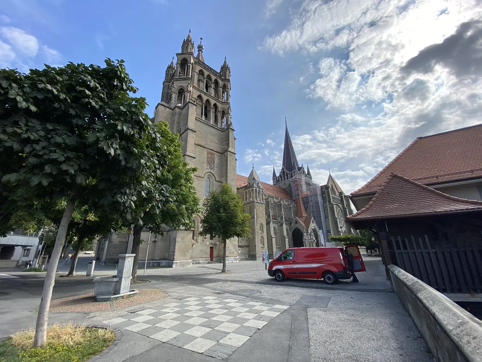 The twin Gothic towers of Lausanne Cathedral rising above a cobbled square where a red van is parked outside a covered market building.