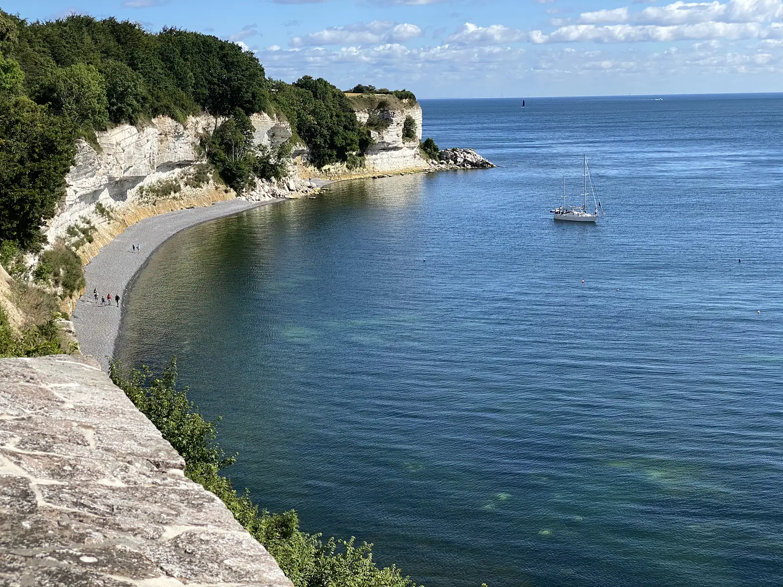 White chalk cliffs curving round a shingle beach above deep blue-green sea, with a yacht at anchor and walkers on the shore.