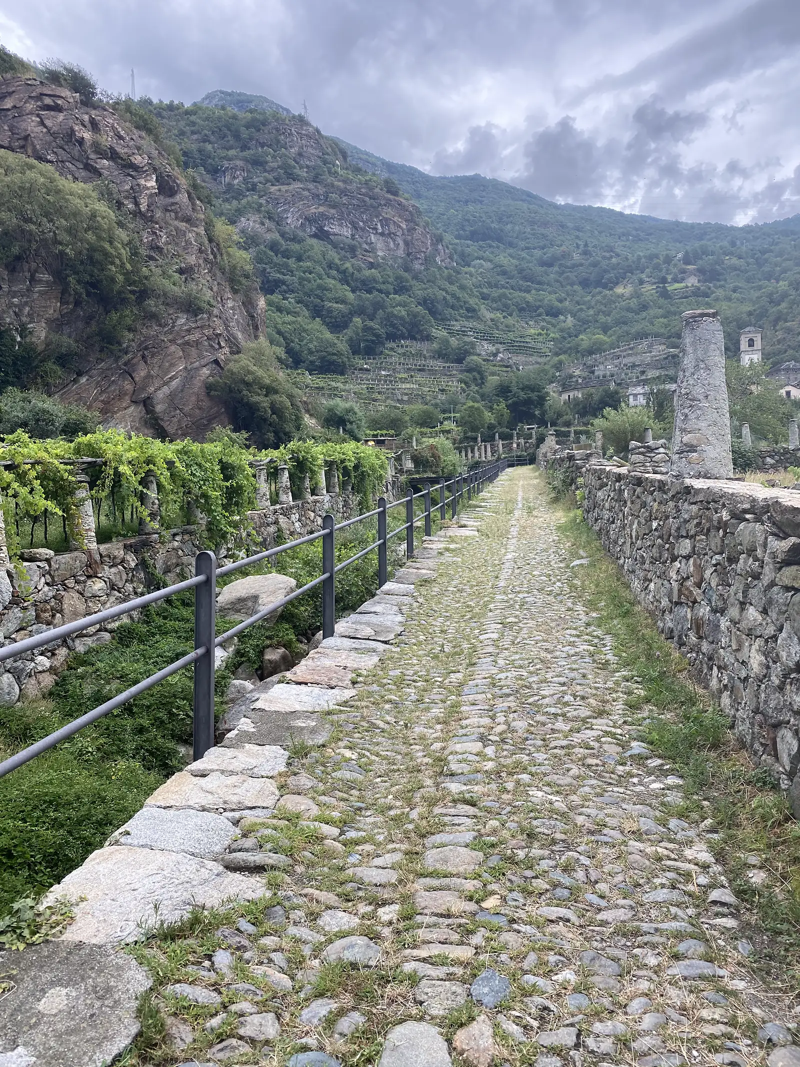 A cobbled lane bordered by dry-stone walls and trellised vines leads towards a village at the foot of a wooded, rocky hillside in the Aosta Valley.