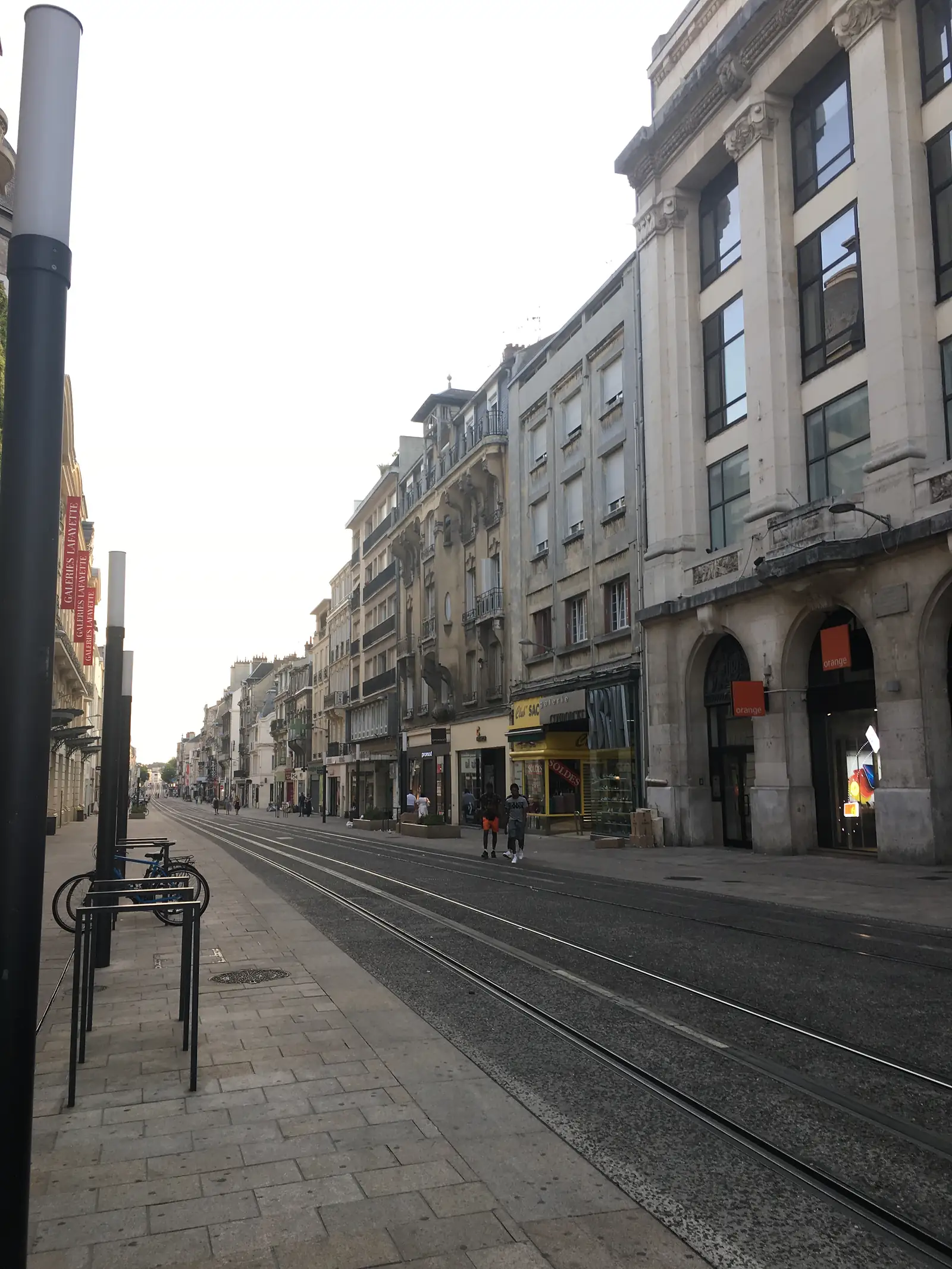 Tram tracks run down a wide pedestrianised shopping street past an Orange phone shop and a Galeries Lafayette store sign.