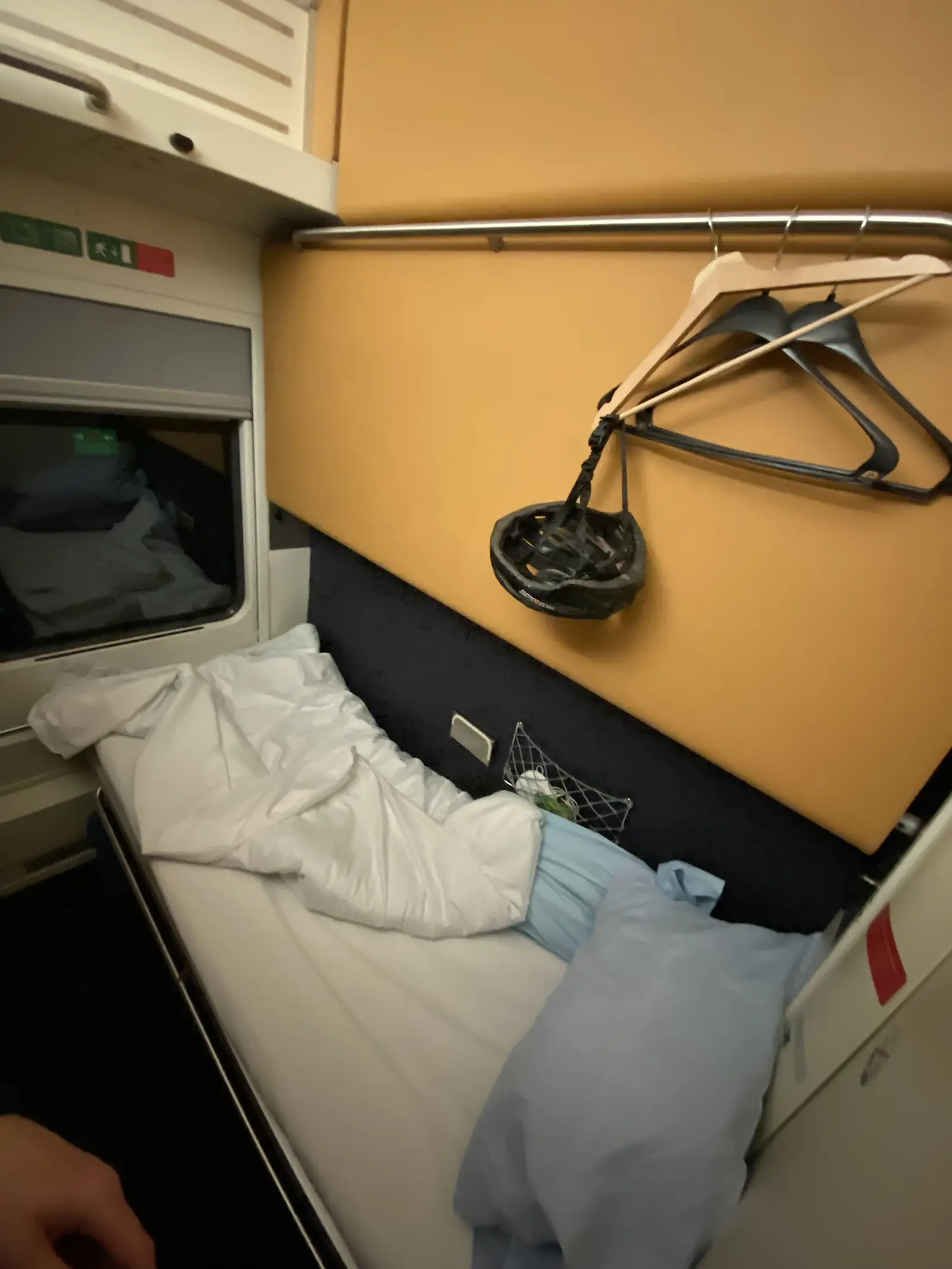 A cycling helmet hangs from a clothes hook above a made-up bed in a train sleeper compartment.