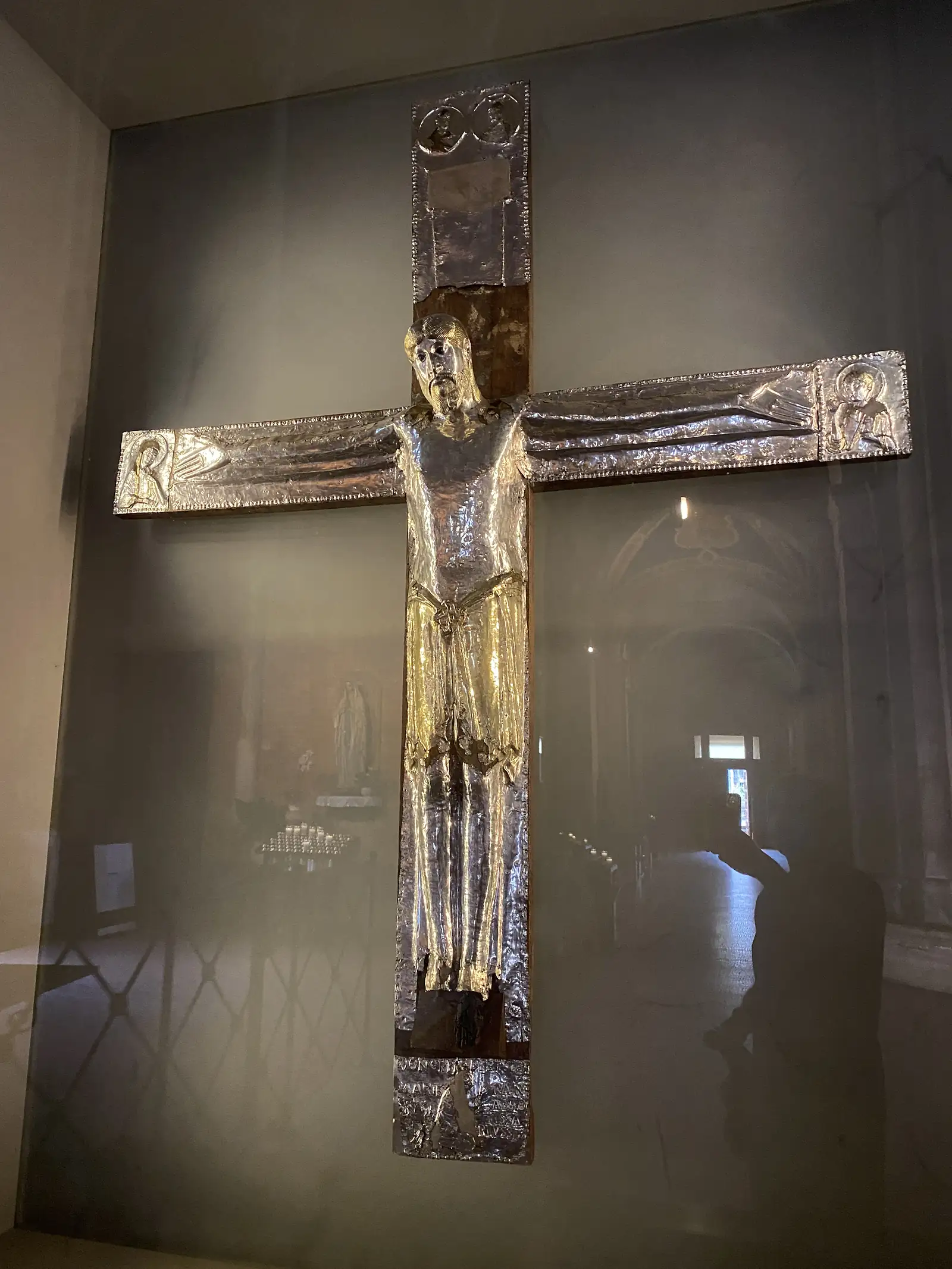 A silver and gold crucifix, its figure of Christ worked in repoussé metal, is displayed in a glass museum case.