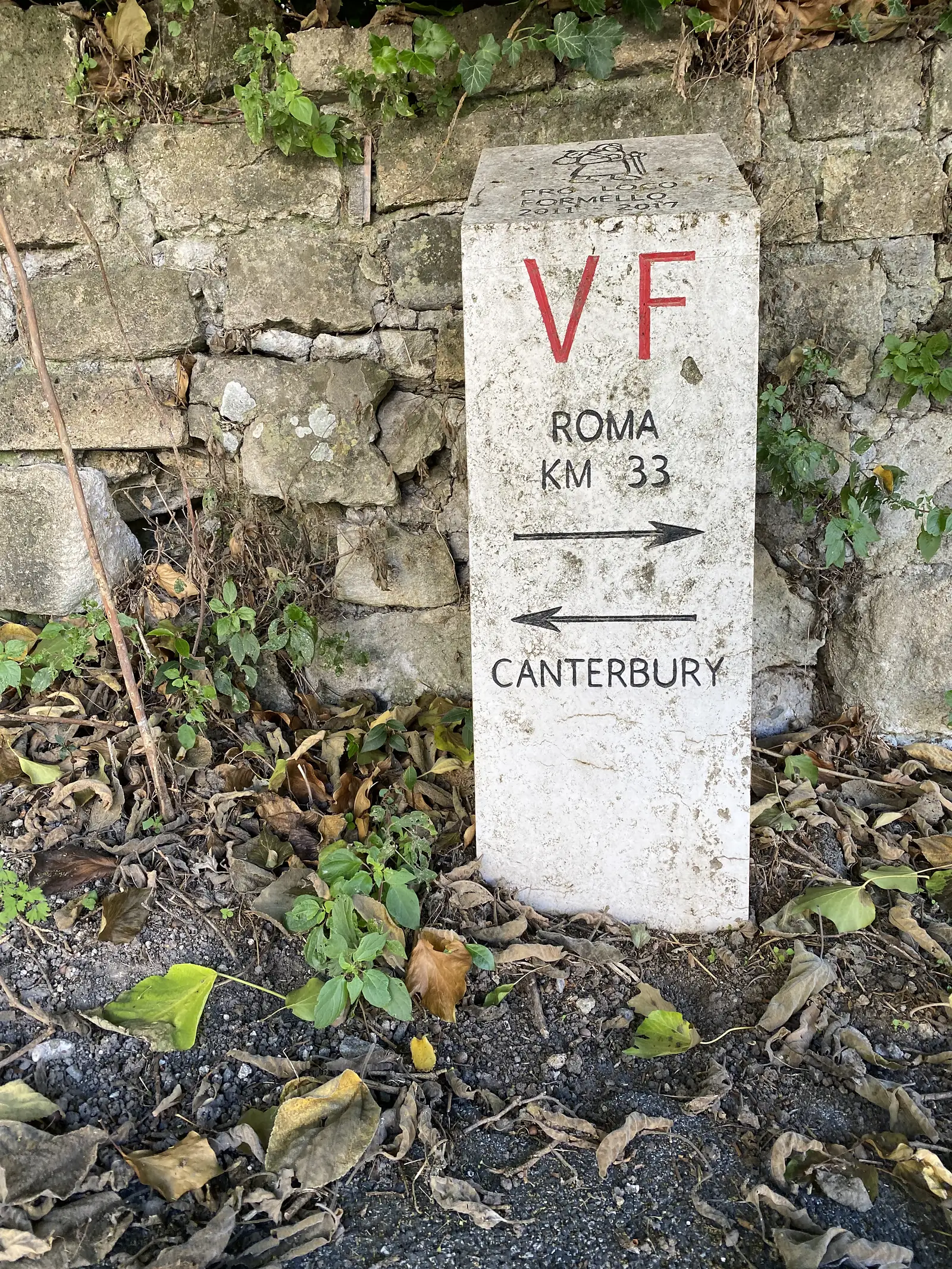 A stone Via Francigena waypost marked "VF, Roma km 33" with arrows pointing onward to Rome and back towards Canterbury, set among weeds on a low wall.