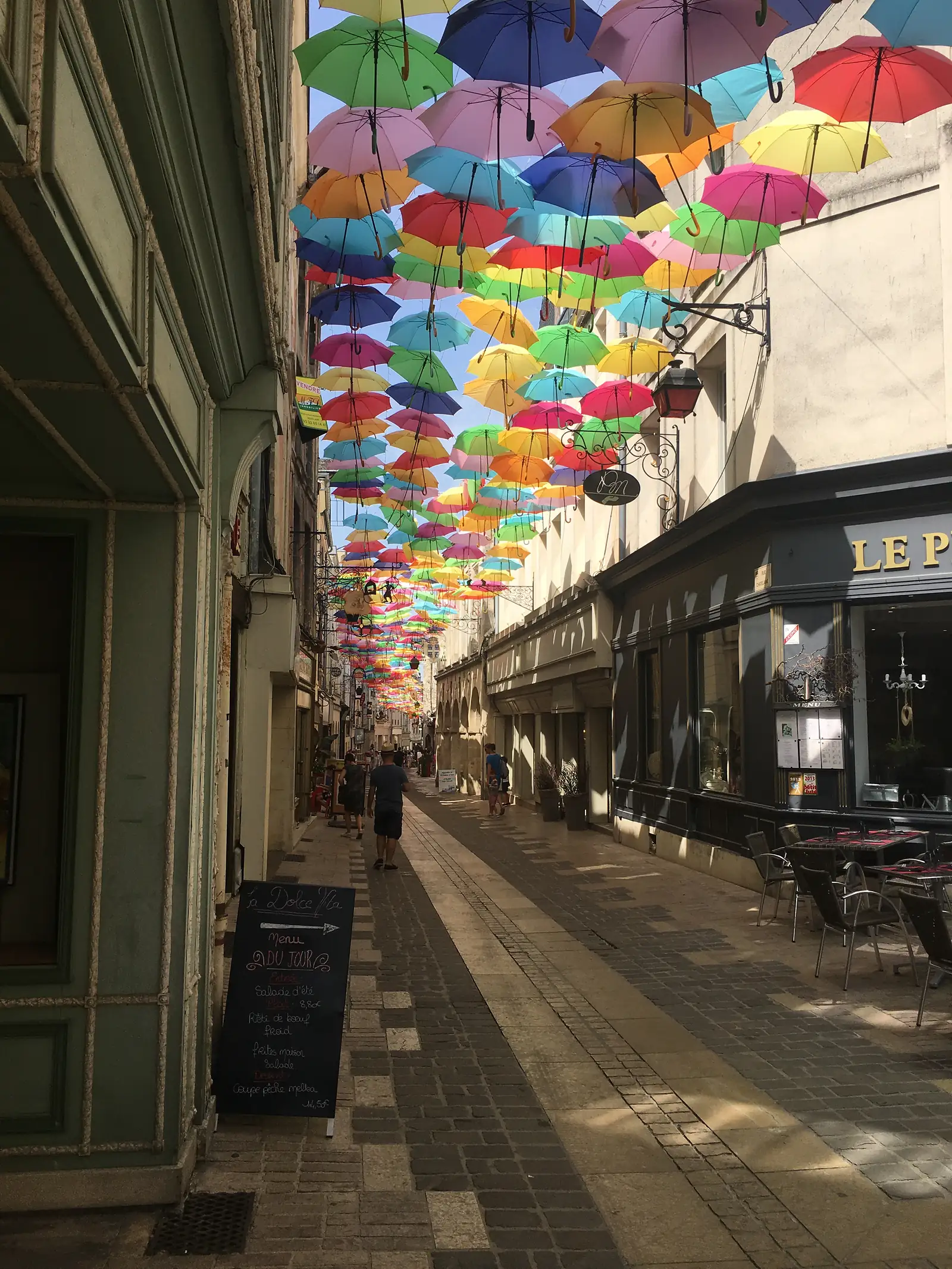 Hundreds of open umbrellas in red, yellow, green, blue and pink hang suspended above a cobbled shopping street, casting coloured light onto the pavement below.